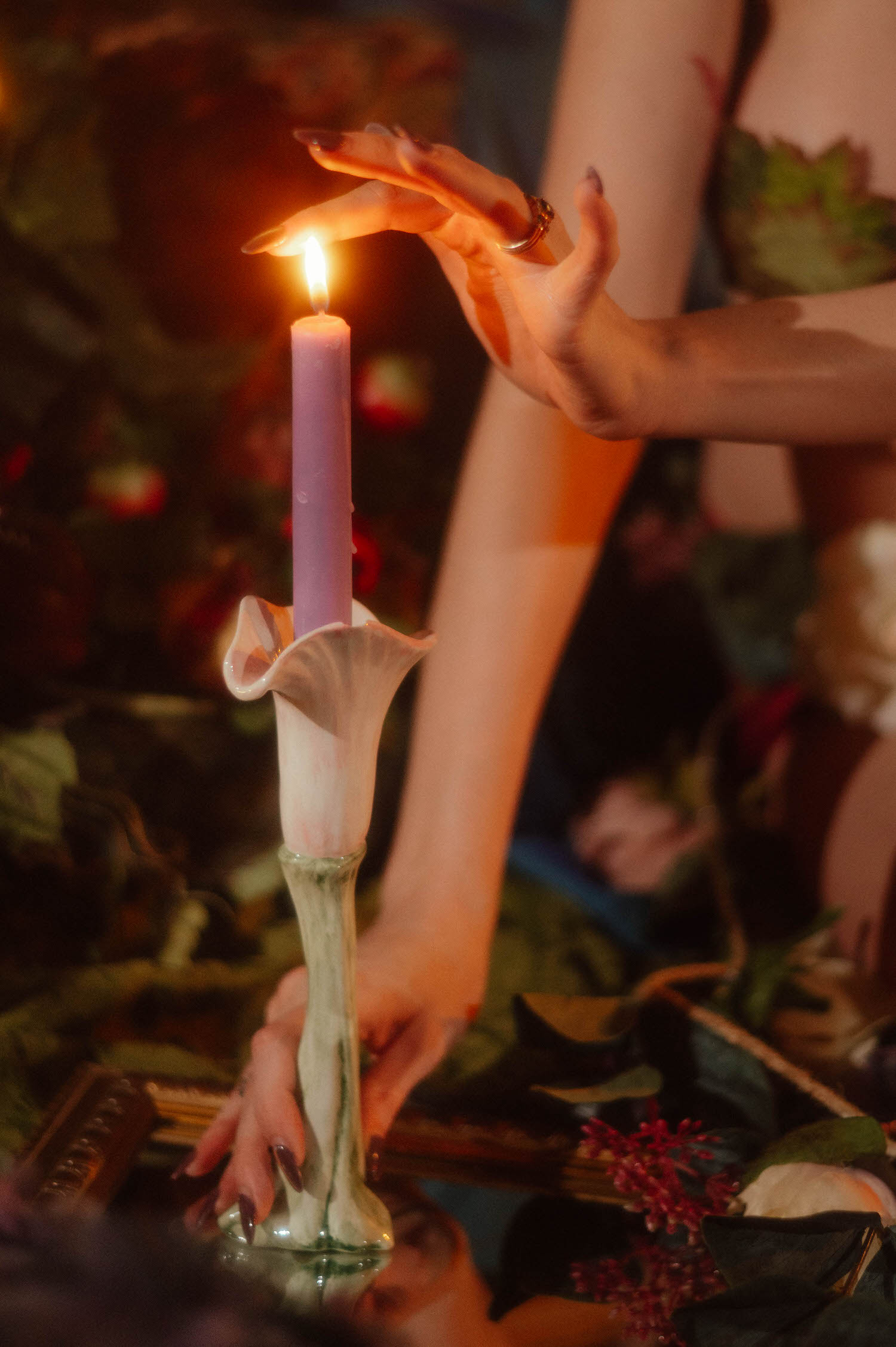 A close up shot shows a hand with dark nails and gold rings hovering over a lit lavender candle in a calla lily shaped holder, surrounded by soft greenery, styled around the whimsigoth aesthetic at Royal Lune Photo.