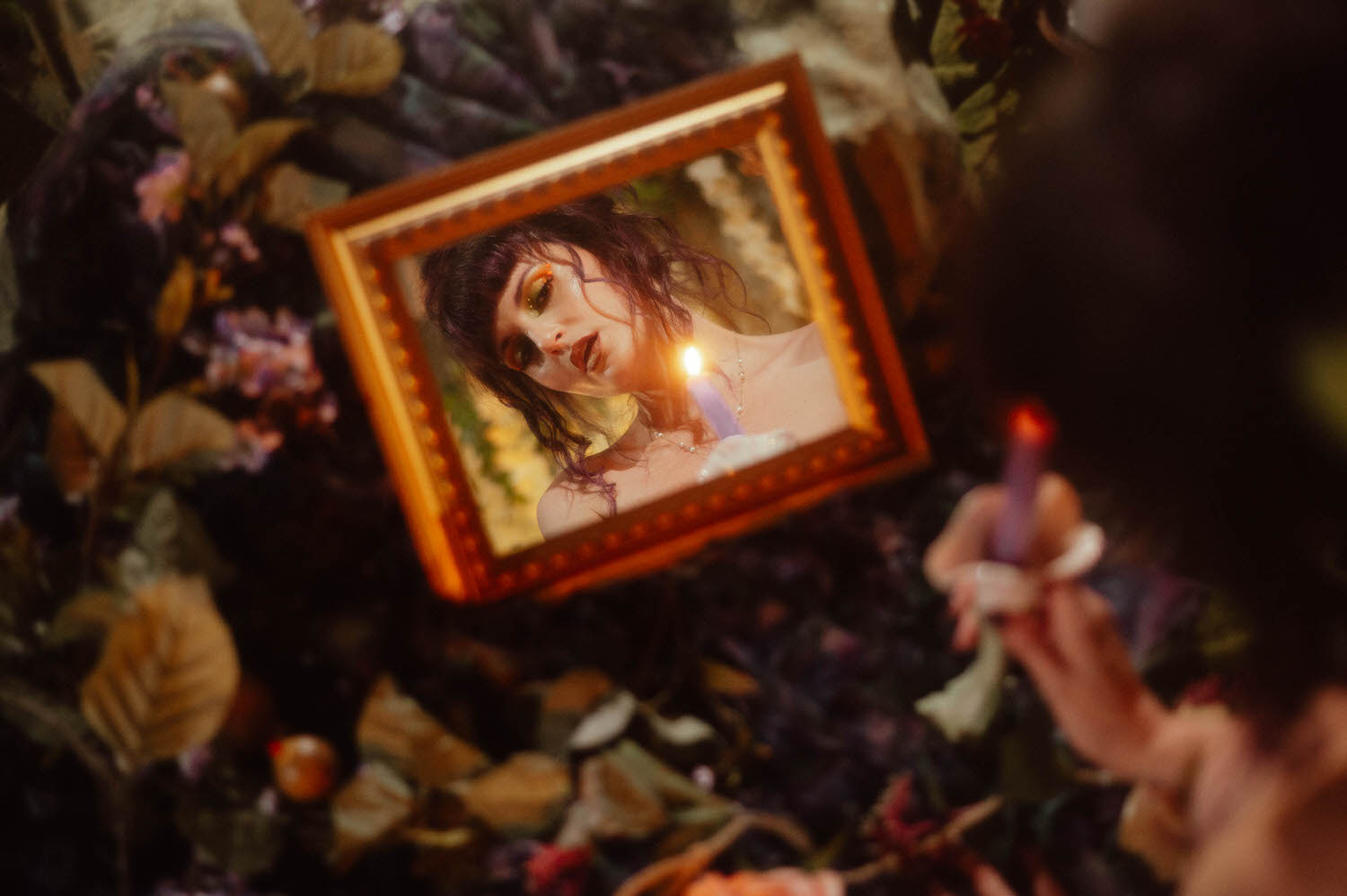 A framed mirror reflects a model's face lit by a single lavender candle, held by a hand with dark manicured nails against a backdrop of dried florals, part of a dark fairytale boudoir photoshoot at Royal Lune Photo.
