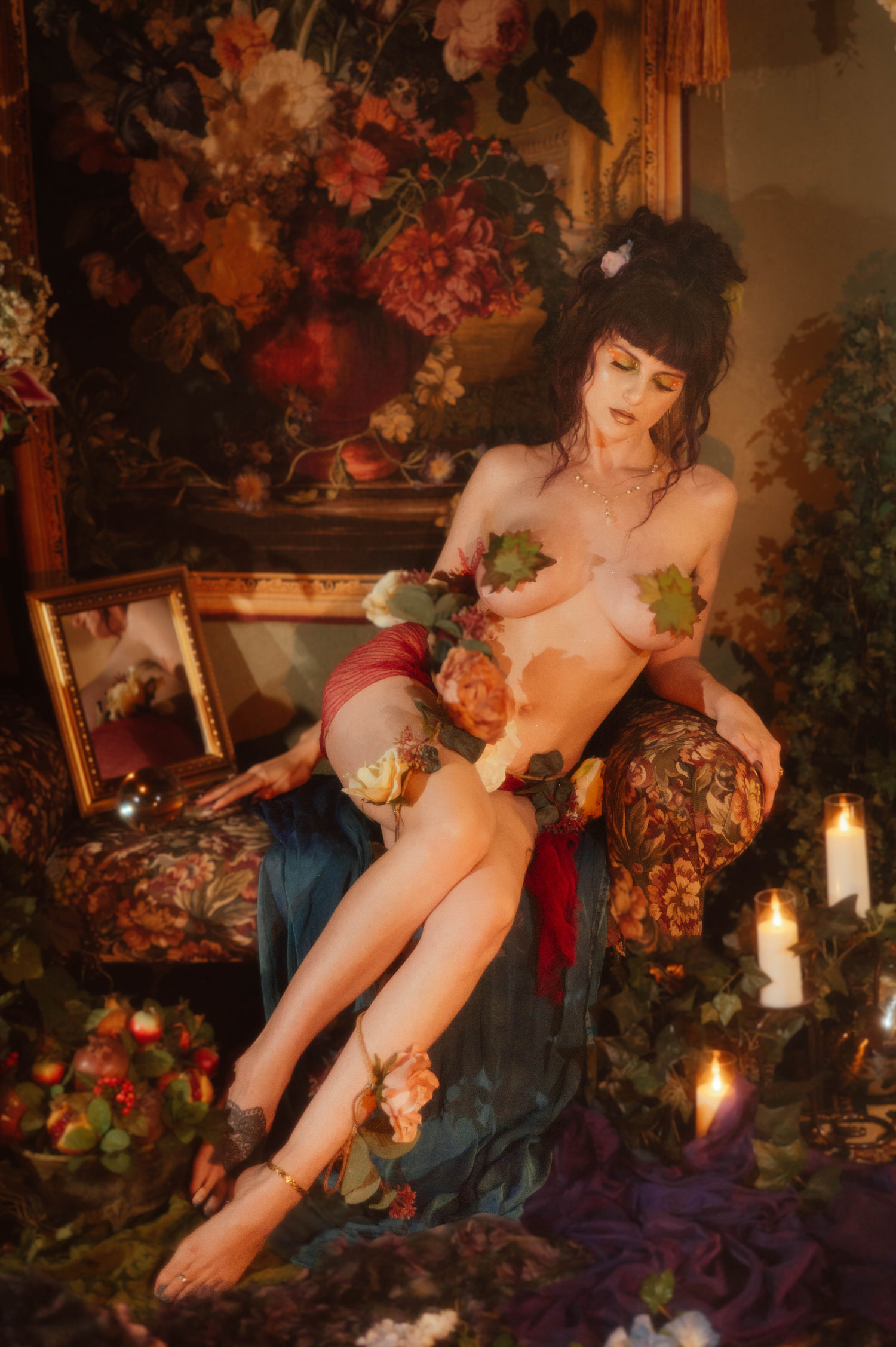 A model reclines in a nude look styled with layered roses and draped red and teal fabric, surrounded by lit candles and a painterly floral backdrop, capturing the final look of a moody boudoir photoshoot at Royal Lune Photo.