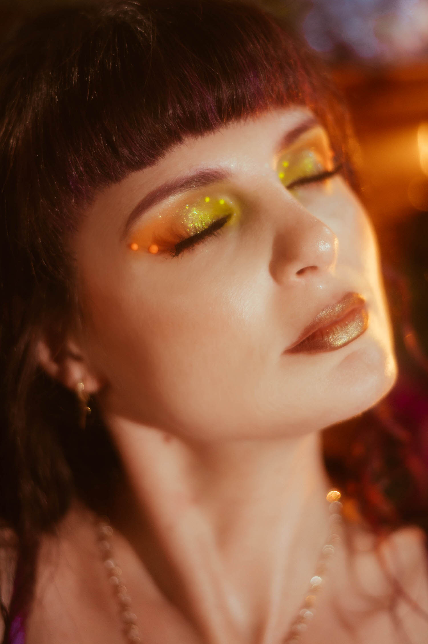 A close up beauty shot features soft orange and gold eyeshadow with a glossy bronze lip and gold hoop earring, lit by warm ambient light during a dark fairytale boudoir photoshoot at Royal Lune Photo.