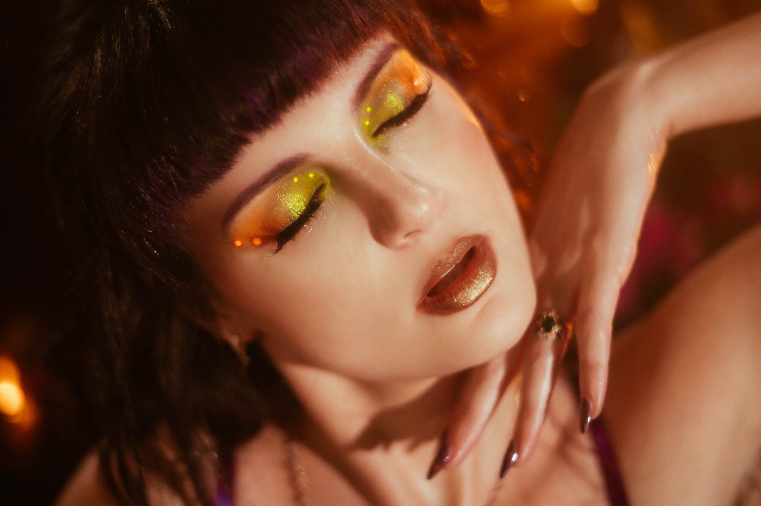 A close up beauty portrait shows a woman with closed eyes wearing shimmering gold eyeshadow and a metallic bronze lipstick, her hand resting near her face with dark manicured nails against warm amber bokeh lighting, showing what to expect at a boudoir photoshoot with professional hair and makeup at Royal Lune Photo.