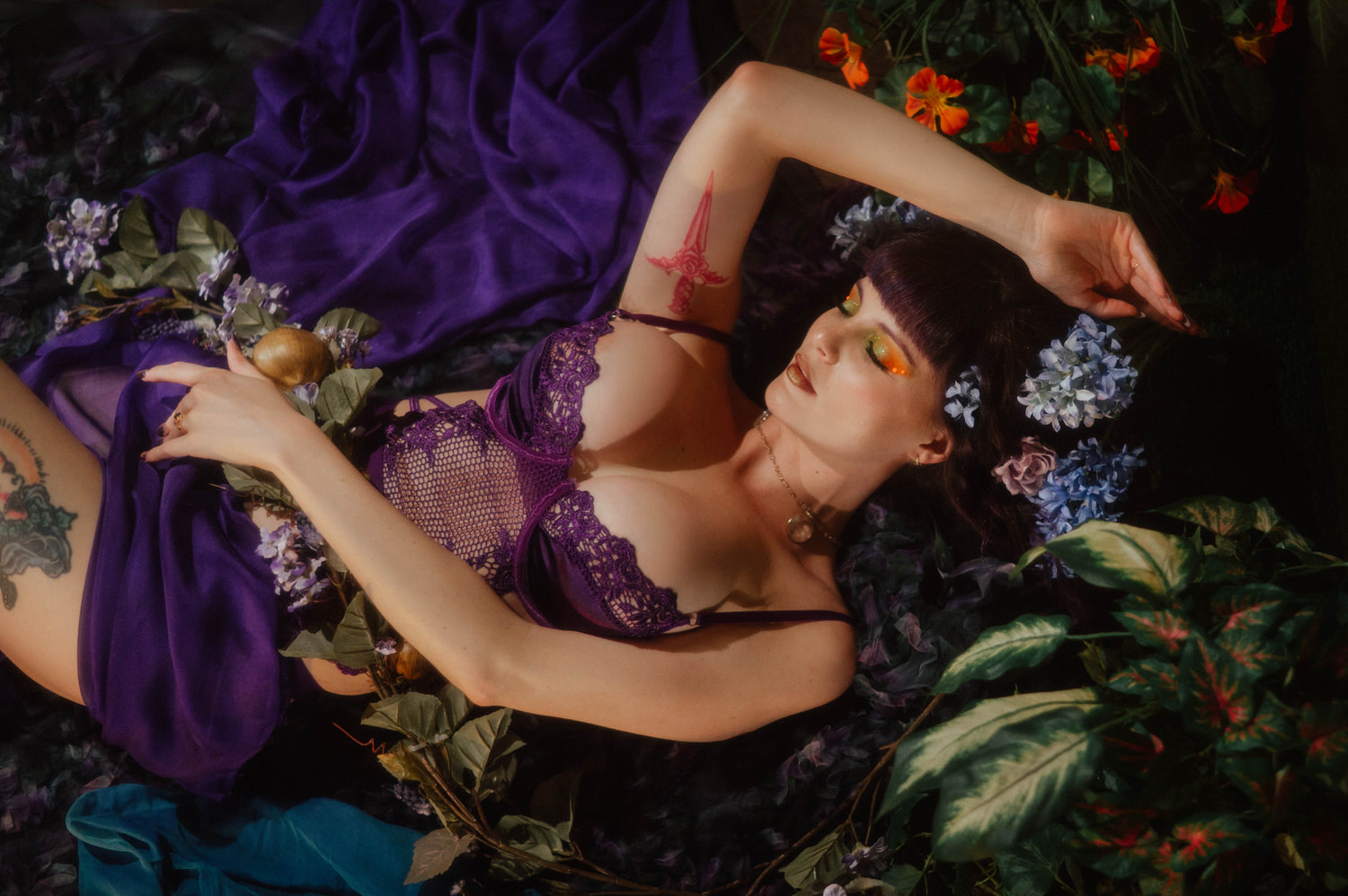 A model lies among purple silk fabric and orange nasturtium flowers in a purple lace bodysuit, with a small red dagger tattoo visible on her inner arm, styled around the whimsigoth aesthetic at Royal Lune Photo.
