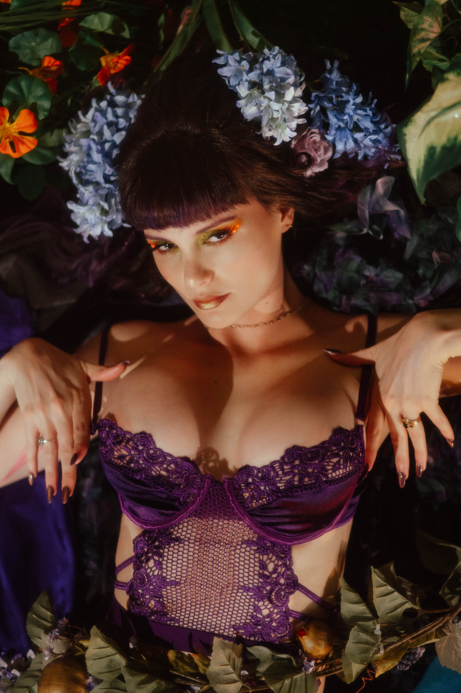 A close up shot shows a model adjusting the straps of her purple lace bodysuit while lying among blue hyacinth and orange florals, styled by a Dallas Fort Worth boudoir photographer at Royal Lune Photo.