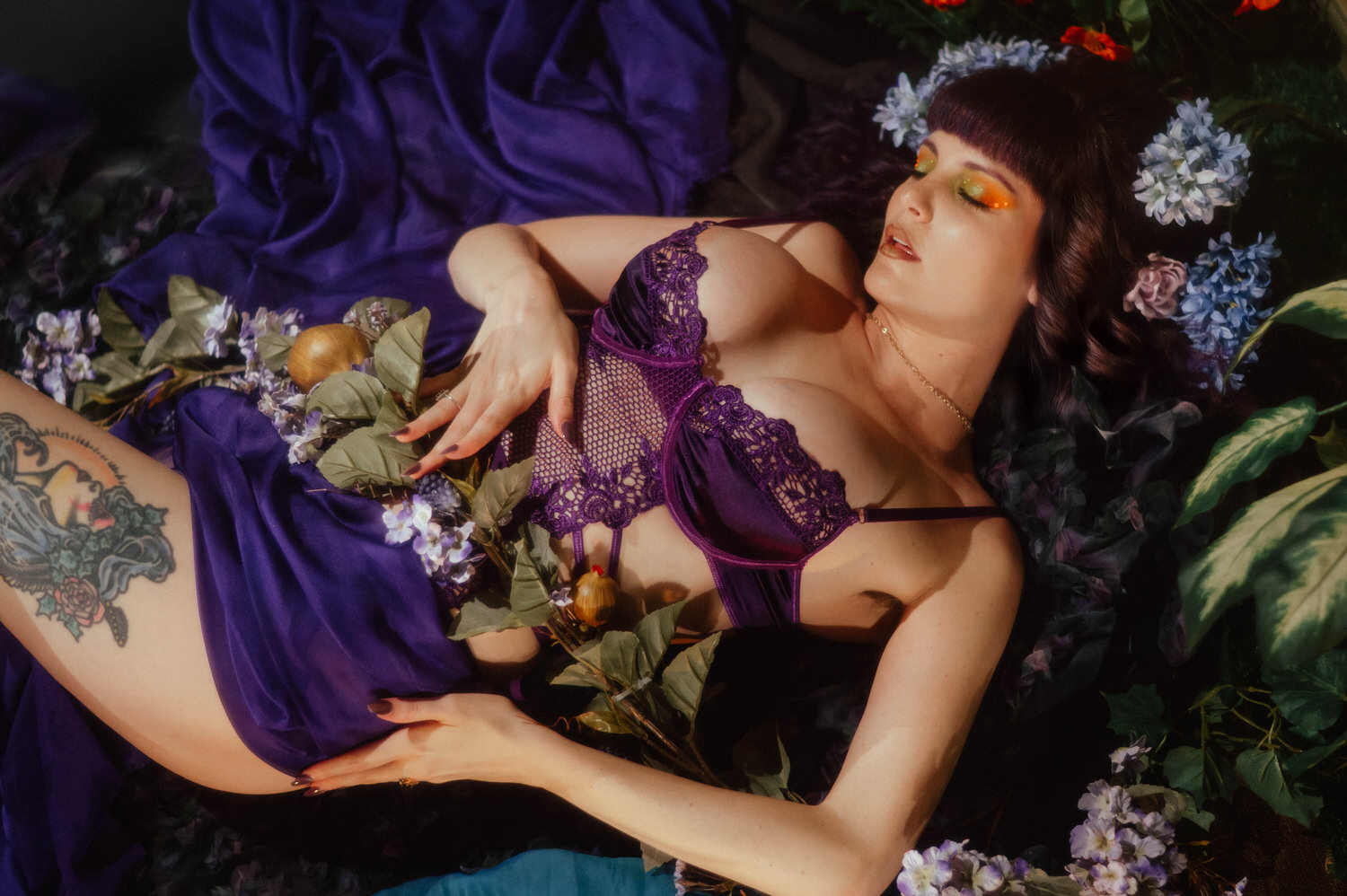A model lies back on purple silk surrounded by lilac blooms and greenery, wearing a purple lace bodysuit with a colorful thigh tattoo catching the warm light, photographed at a Dallas Fort Worth photo studio for Royal Lune Photo.