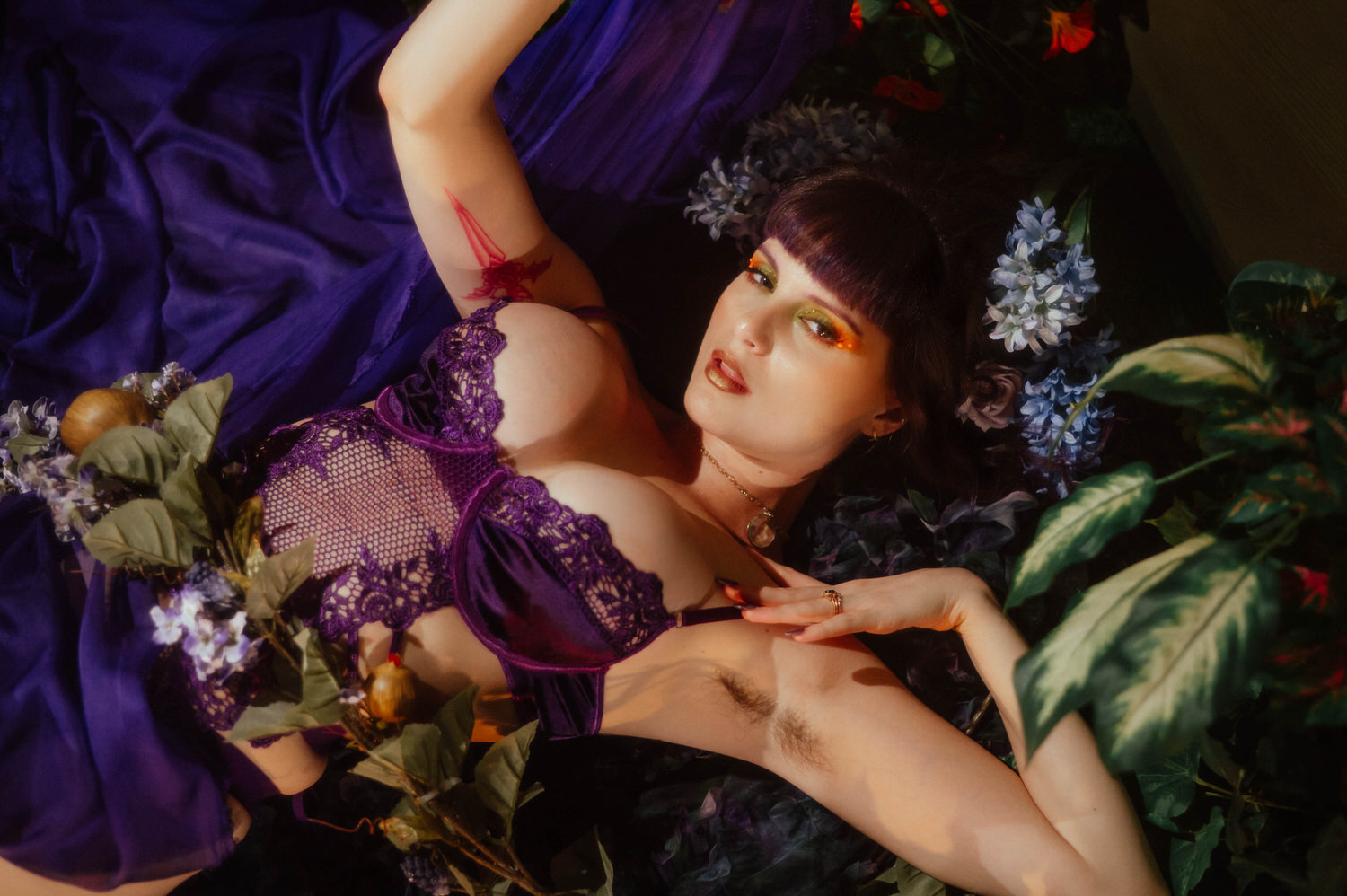 A model lies among purple silk and greenery in a purple lace bodysuit, looking directly at the camera with gold and green eyeshadow catching warm ambient light in a dark fairytale boudoir photoshoot at Royal Lune Photo.
