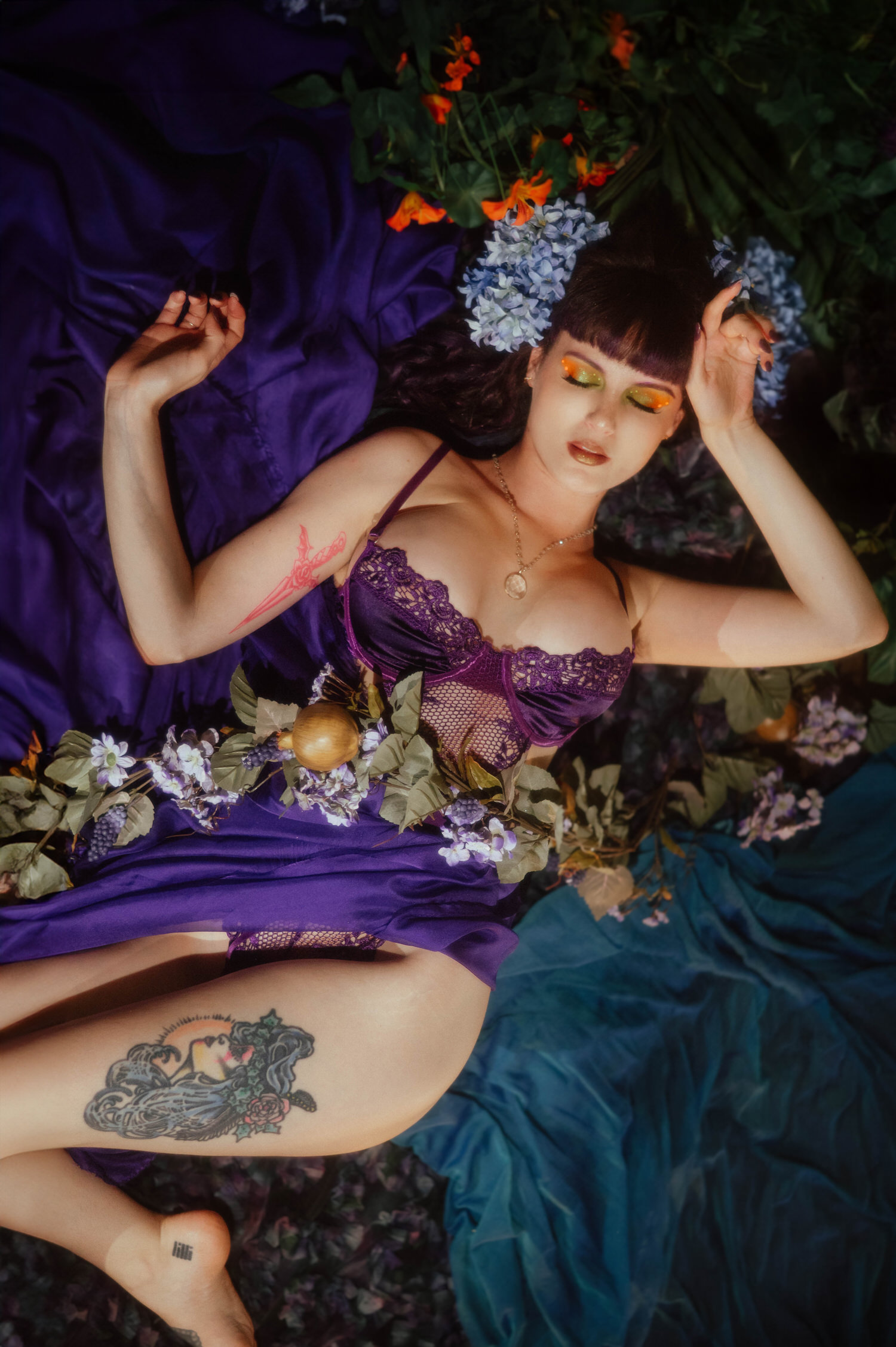 A model lies back among purple silk and hydrangea blooms in a purple lace bodysuit, revealing a detailed color tattoo on her thigh under soft warm lighting, part of a whimsigoth aesthetic gallery at Royal Lune Photo.