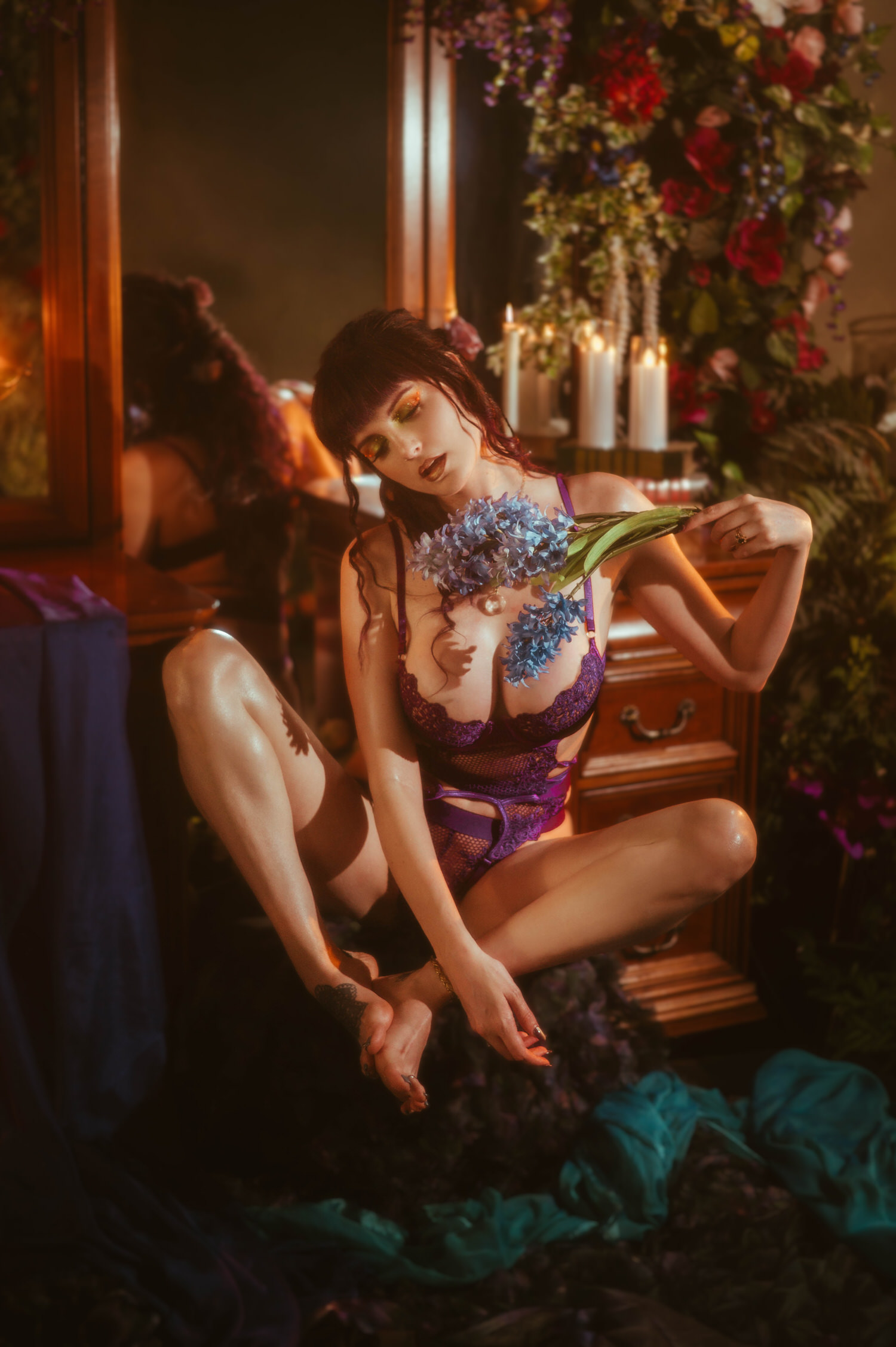A model sits on the floor during a moody boudoir photoshoot, wearing a purple lace bodysuit and holding blue hyacinth against her chest in front of a candlelit antique mirror with a floral arch, styled at Royal Lune Photo.