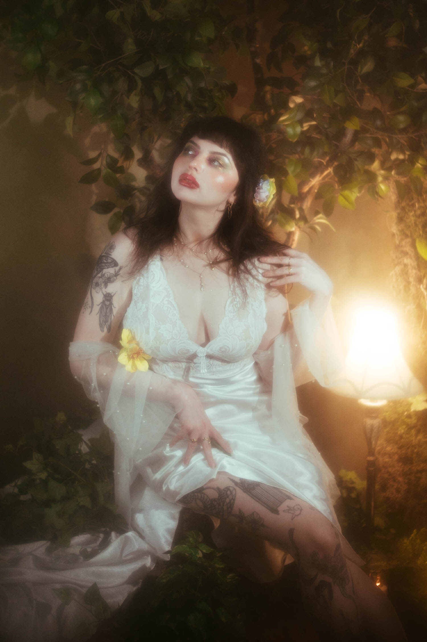 A bride reclines with a yellow flower tucked at her bodice, her tattooed leg extended and lit by warm moody boudoir lighting from a nearby lamp inside Royal Lune's vintage photo studio, Royal Lune Photo.