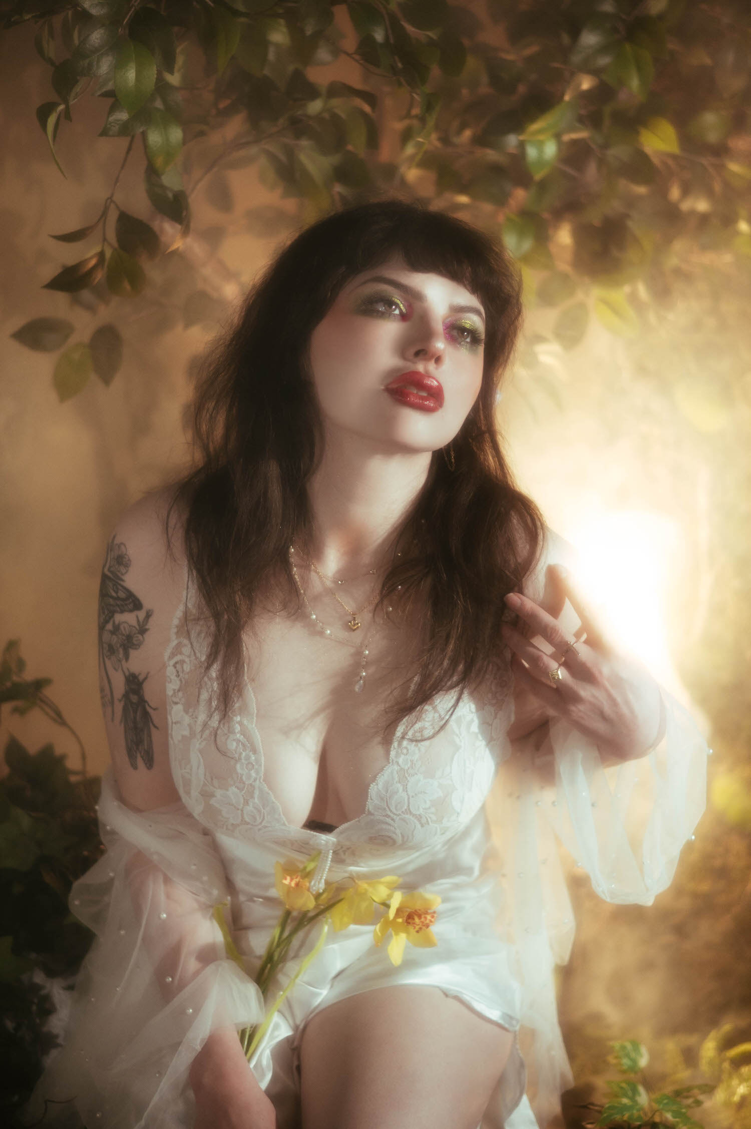 A bride holds her hand up toward a warm light source, yellow flowers resting at her waist and soft fog machine boudoir photography haze glowing around her tattooed arm, Royal Lune Photo.