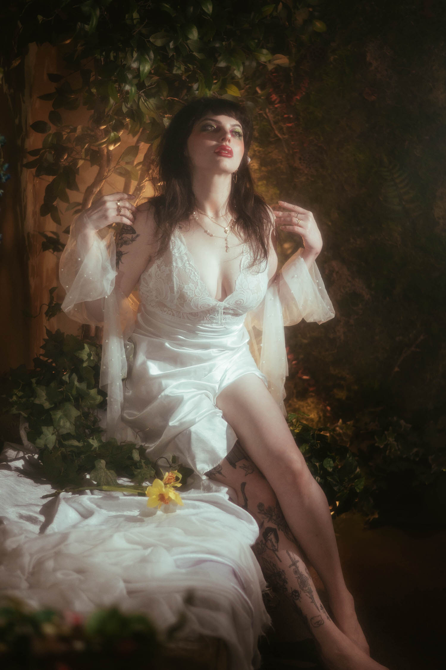 A bride sits with her legs extended and sheer pearl beaded sleeves raised at her sides, a single yellow flower resting on white fabric nearby during this moody boudoir lighting portrait at Royal Lune Studio, Royal Lune Photo.