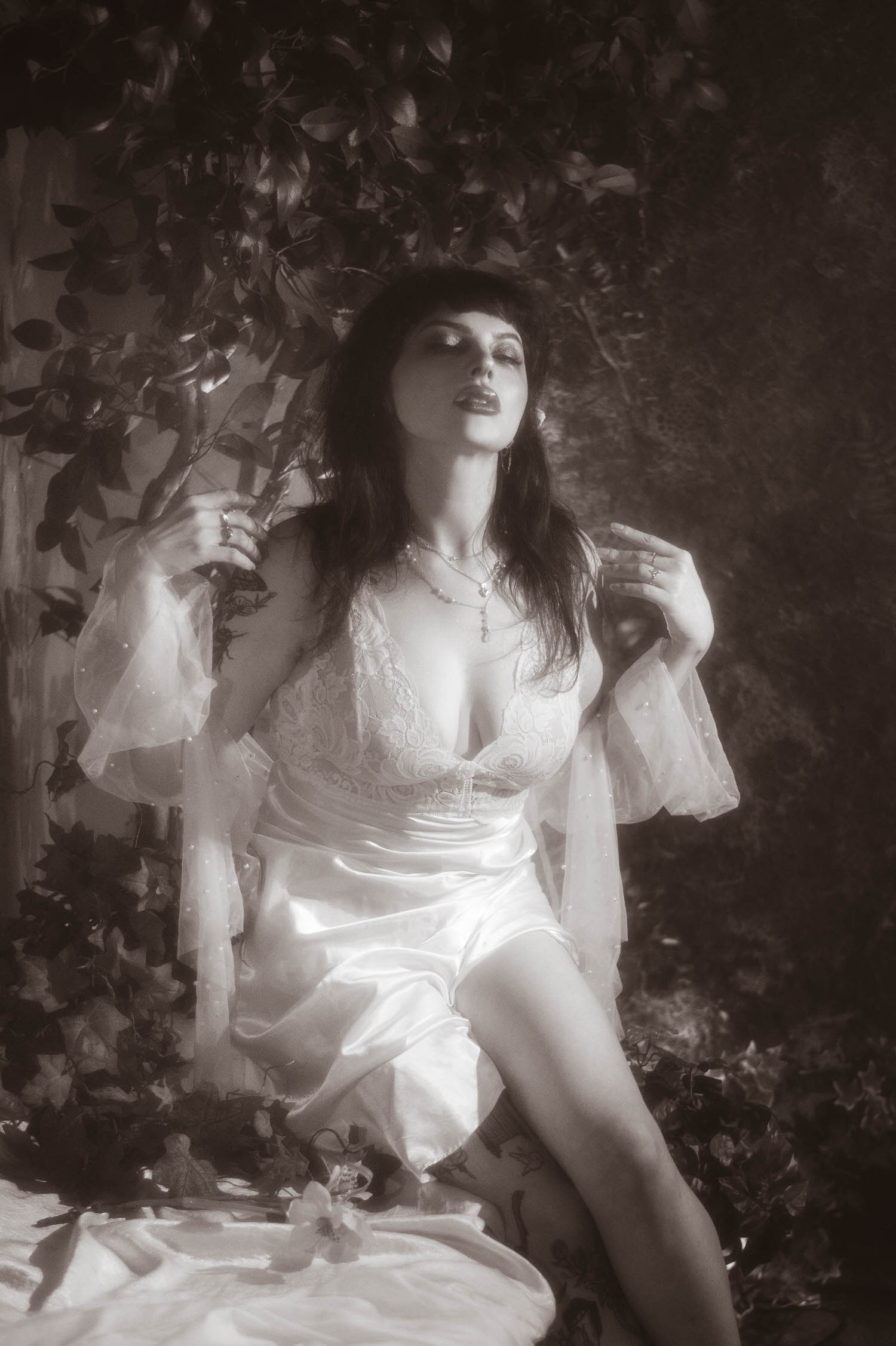 A black and white portrait shows a bride in a vintage nightgown boudoir look with her eyes closed and sheer pearl beaded sleeves raised, deep shadow and soft highlights shaping this bridal boudoir photography image at Royal Lune Studio, Royal Lune Photo.