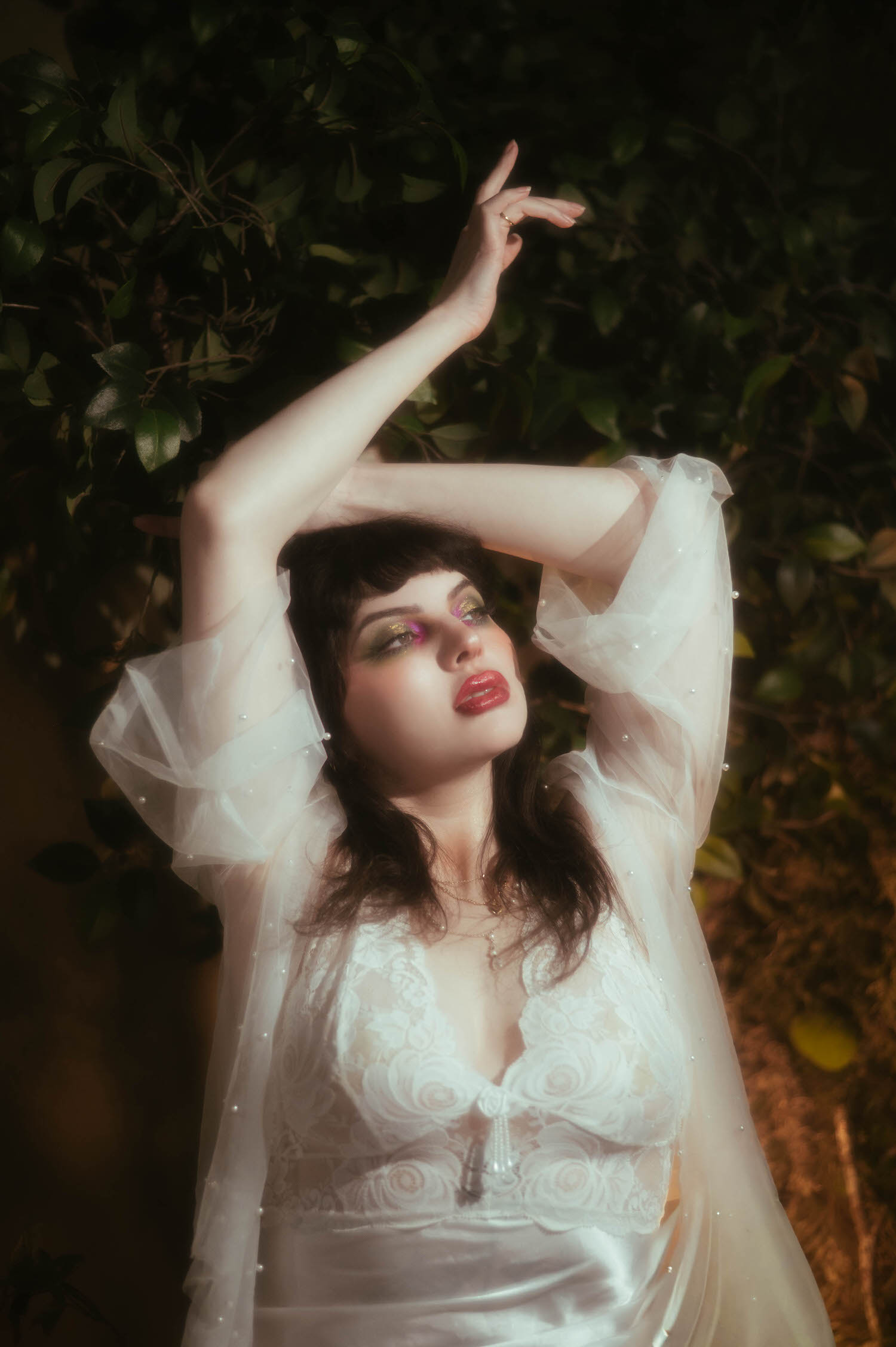 A bride raises both arms overhead in a vintage nightgown boudoir look, her sheer pearl beaded sleeves catching warm light against a dark greenery backdrop as a DFW bridal boudoir photographer captures the dramatic pose, Royal Lune Photo.