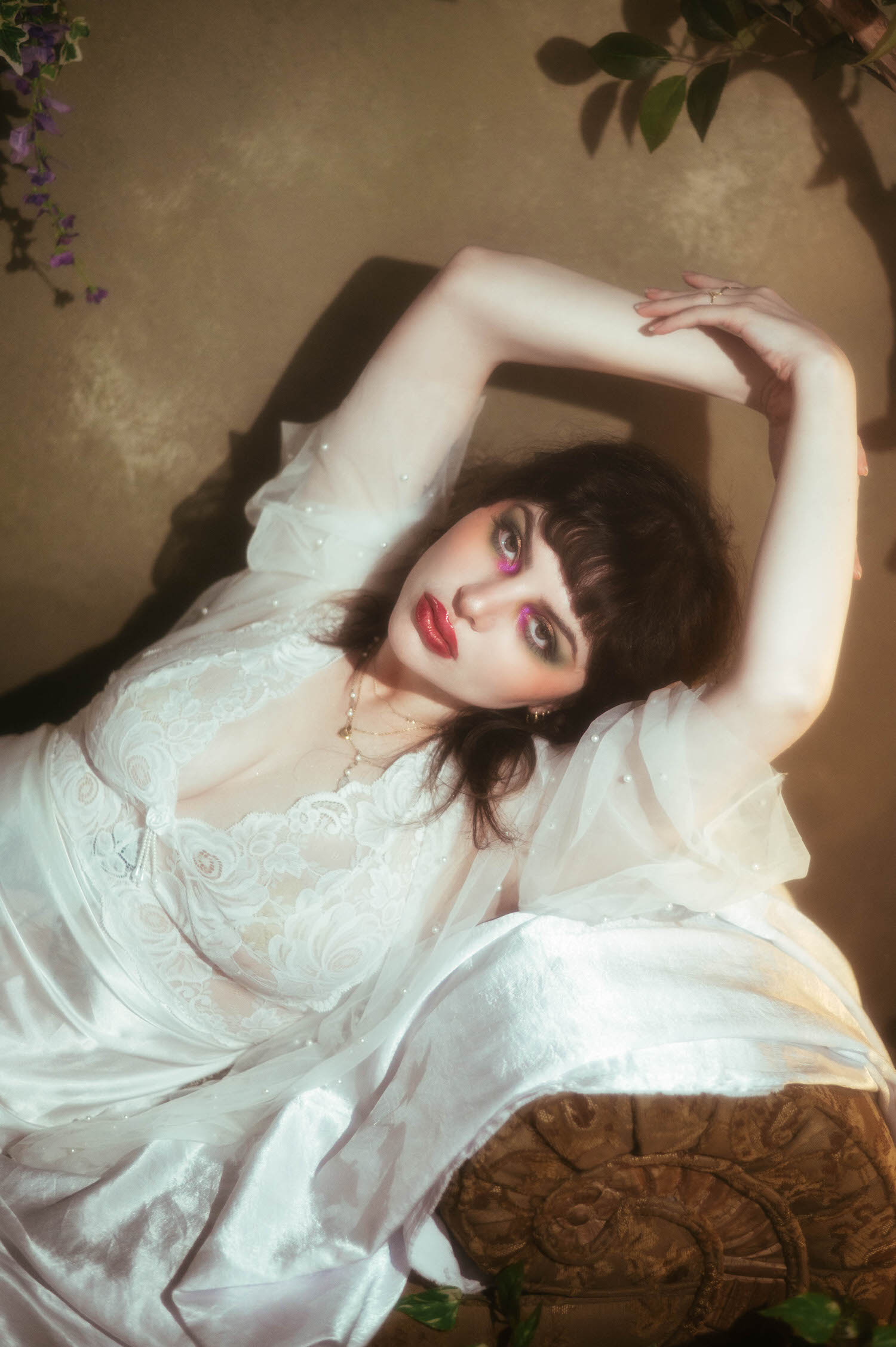 An overhead angle captures a bride lying back with her arms crossed above her head, sheer pearl beaded sleeves draping across the lounge as moody boudoir lighting warms her lace nightgown and bold red lips, Royal Lune Photo.
