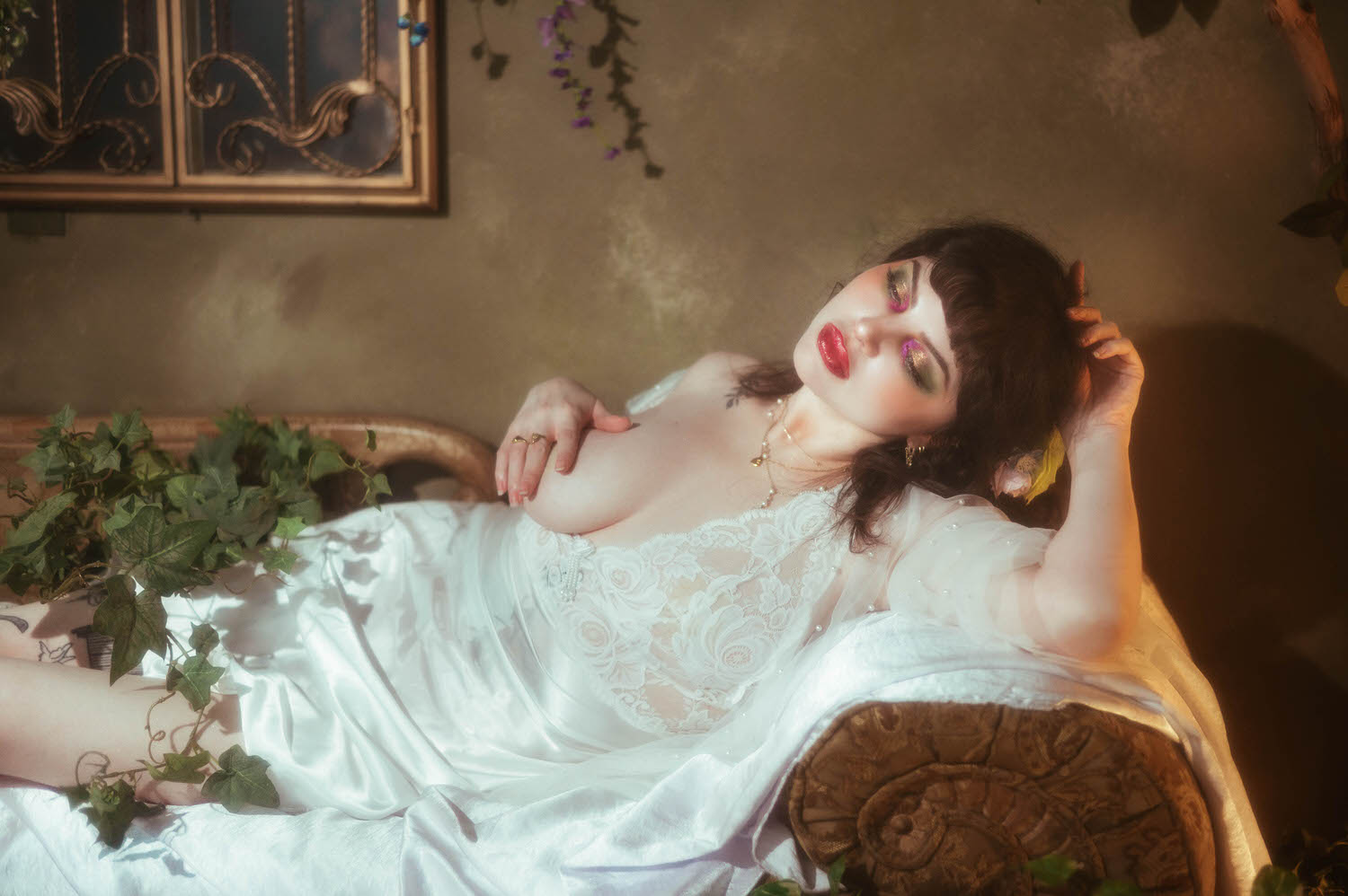 A bride reclines on an antique lounge in a vintage nightgown boudoir look with her hand resting in her hair, trailing ivy spilling across the cushions beside her tattooed leg during this bridal boudoir photography session, Royal Lune Photo.