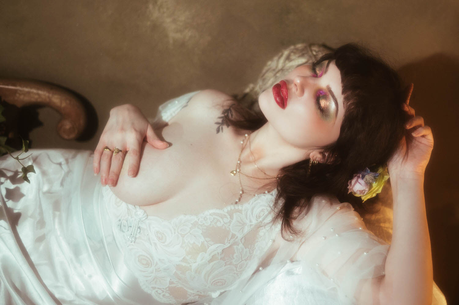 A close up portrait shows a bride reclining with a small pink and yellow flower tucked into her dark hair, her hand resting near her head as warm moody boudoir lighting highlights her lace nightgown and pearl necklace, Royal Lune Photo.