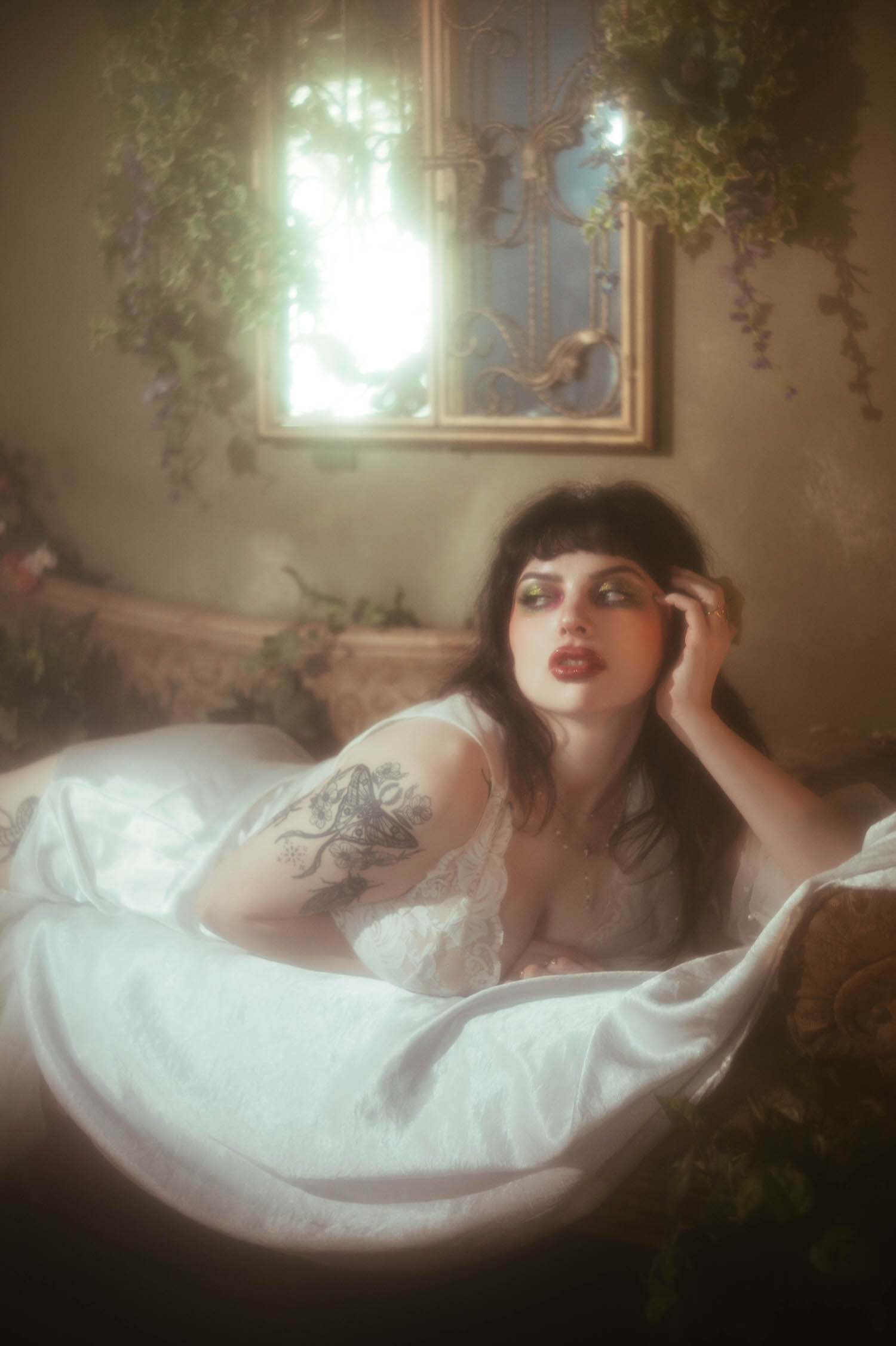 A bride leans forward on an antique lounge in a vintage nightgown boudoir look, bright window light glowing behind a gold mirror sconce and trailing greenery as a DFW bridal boudoir photographer captures her tattooed arm and soft glam, Royal Lune Photo.