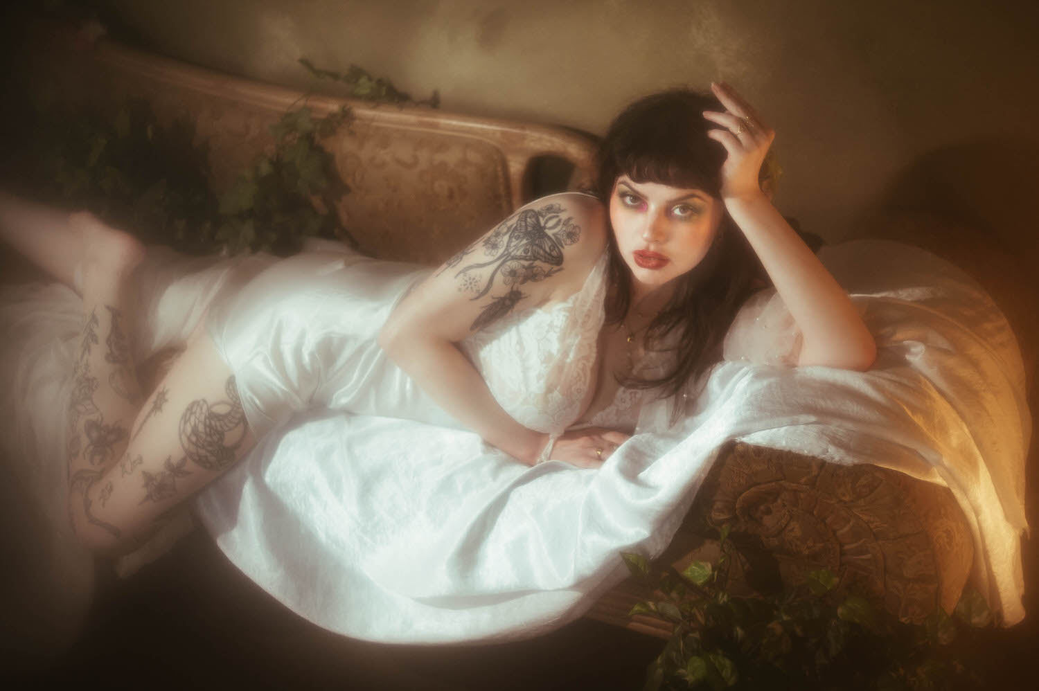 A bride reclines lengthwise across an antique tufted lounge in a vintage nightgown boudoir look, her tattooed leg extended and one hand resting in her hair as warm light and trailing ivy frame this bridal boudoir photography session at Royal Lune Studio, Royal Lune Photo.