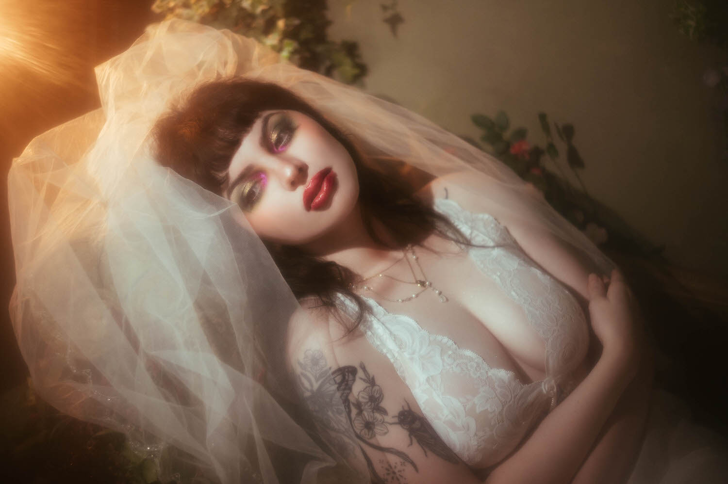 An overhead angle captures a bride lying back with her veil pooling behind her head, her tattooed chest and lace nightgown lit by warm moody boudoir lighting and soft floral tones, Royal Lune Photo.
