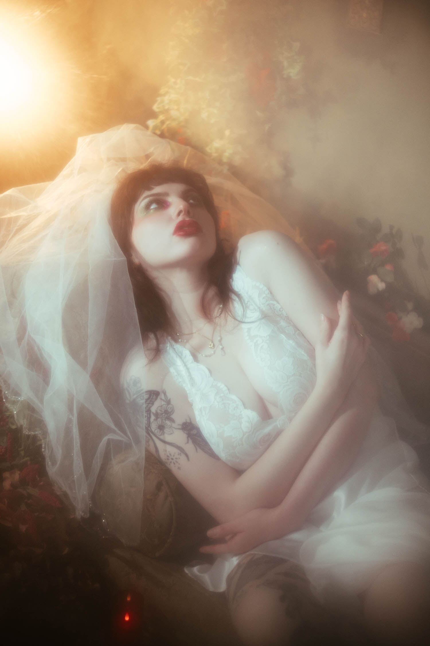 An overhead angle shows a bride looking upward with her arms crossed over her lace nightgown, her veil catching golden light against a blurred floral backdrop inside Royal Lune's vintage photo studio, Royal Lune Photo.