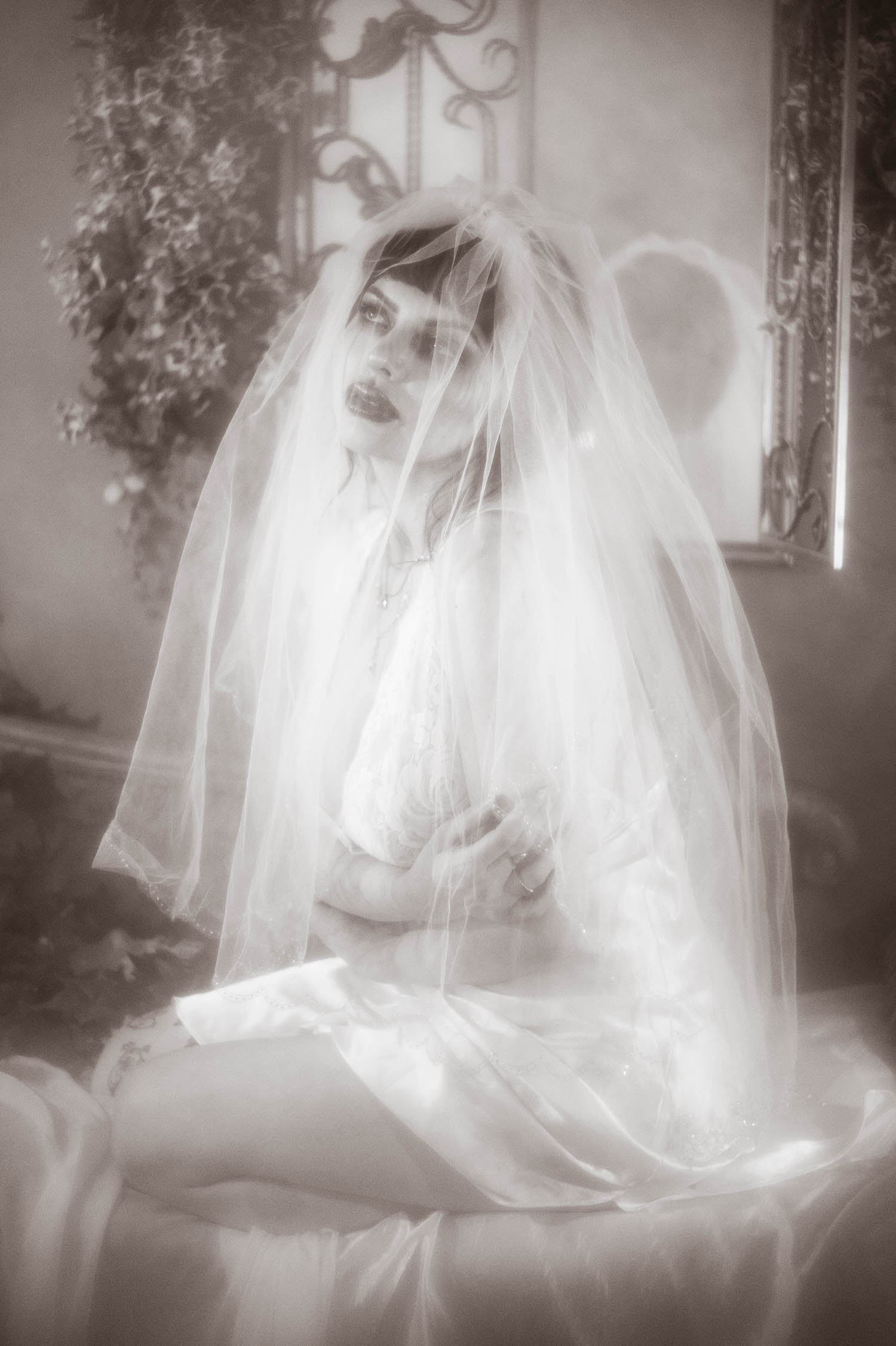 A black and white portrait shows a bride with a sheer veil draped fully over her lace nightgown, her hands folded gently in her lap against a softly lit iron and floral backdrop, Royal Lune Photo.