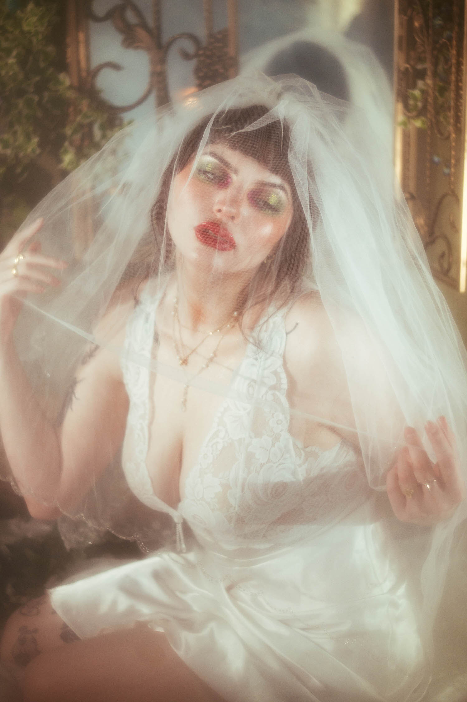 A bride lifts a sheer tulle veil forward over her face with both hands, bold red lipstick and shimmering eyeshadow visible through the fabric as fog machine boudoir photography haze softens the antique iron backdrop, Royal Lune Photo.
