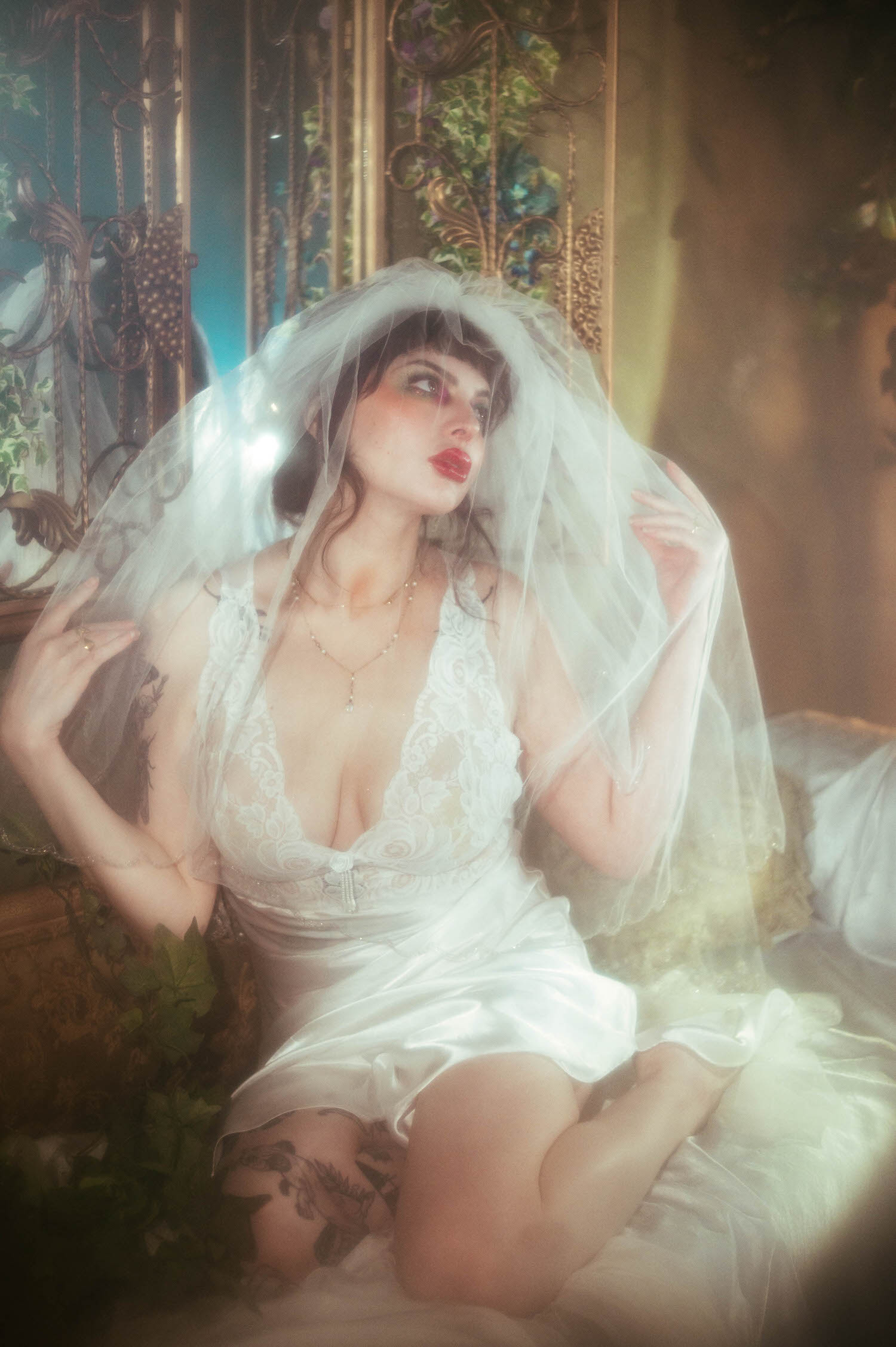 A bride lifts her sheer veil with both hands while seated with her tattooed leg crossed, bold red lips and soft glam visible beneath the fabric during this DFW bridal boudoir photographer session, Royal Lune Photo.