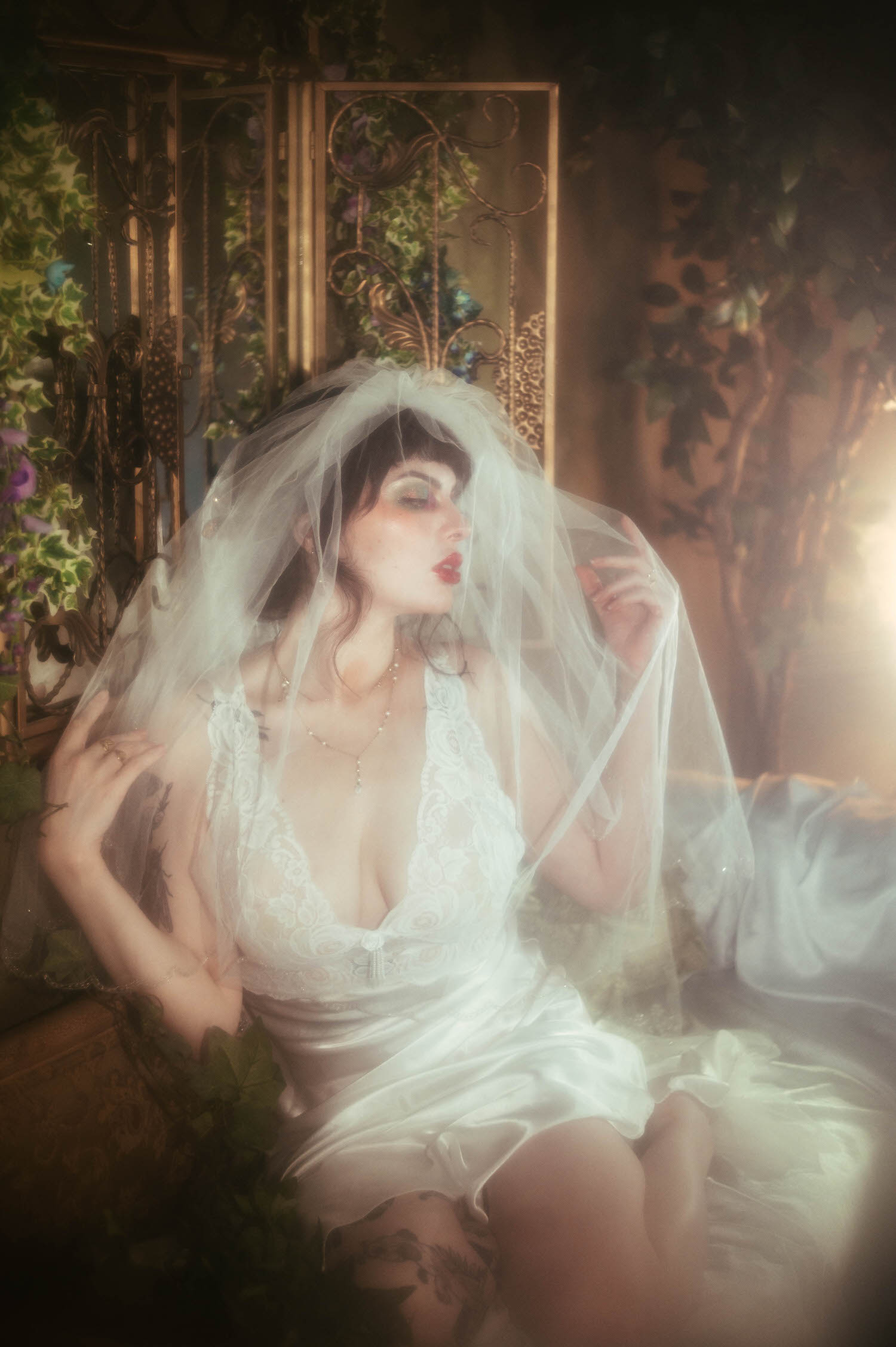 A bride pulls her veil to one side in profile, her lace nightgown and pearl necklace catching warm golden light against an ivy draped iron screen inside Royal Lune's vintage photo studio, Royal Lune Photo.