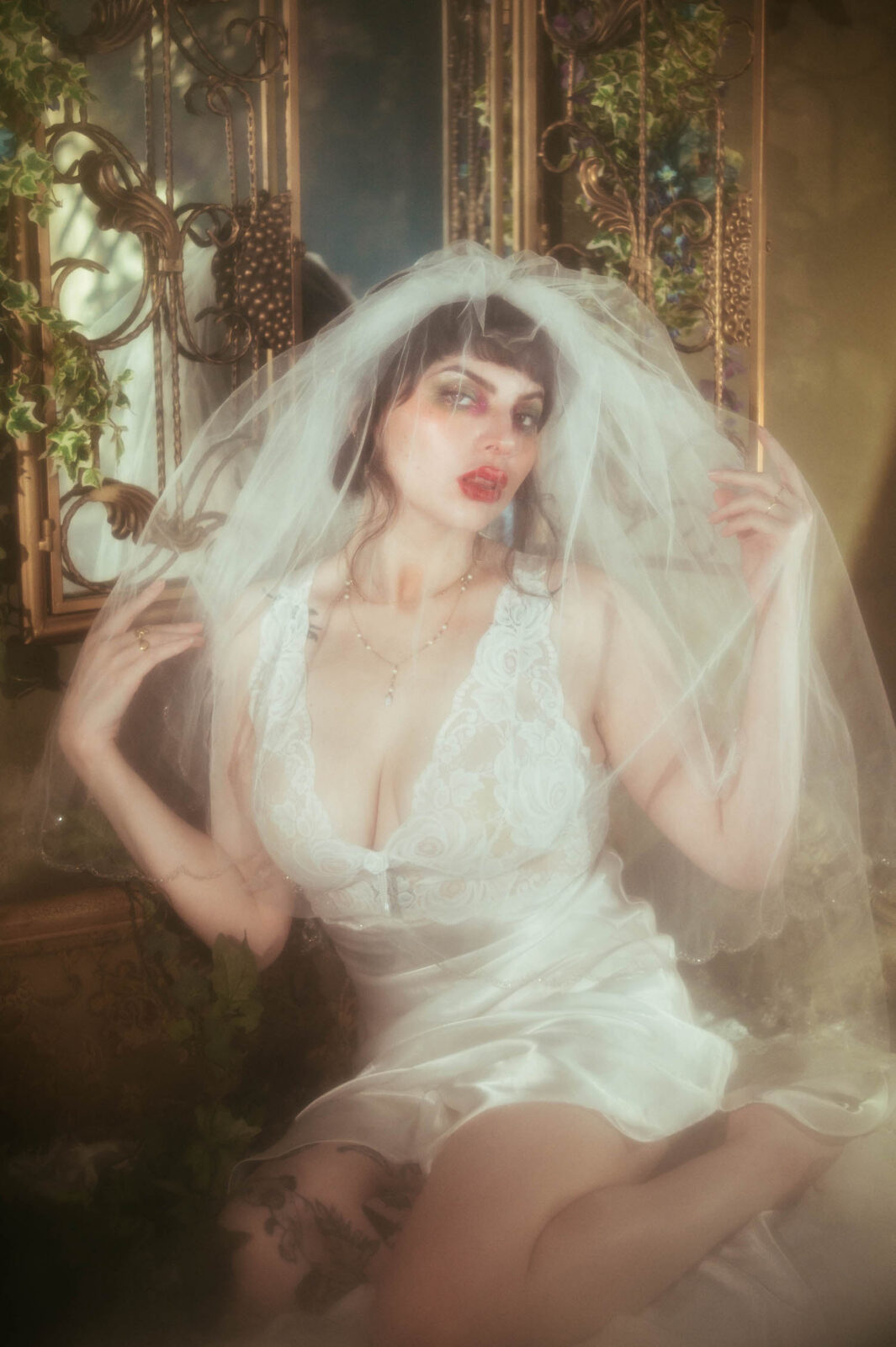 A bride in a vintage nightgown boudoir look lifts a sheer tulle veil overhead while sitting inside a vintage photo studio in front of an antique gold mirror screen, her tongue peeking out playfully beneath bold red lipstick and warm golden light, Royal Lune Photo.