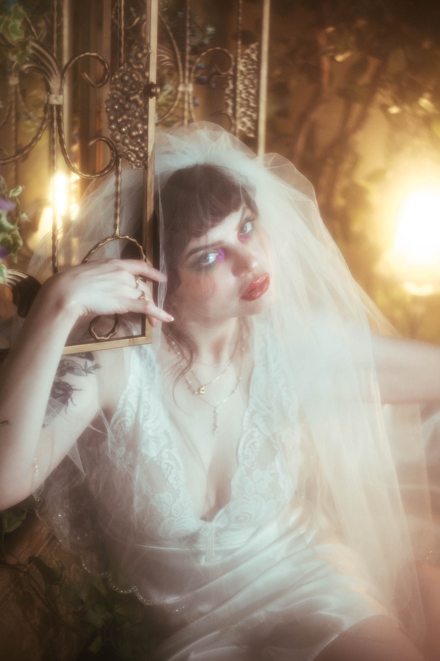 A bride leans her head against an antique iron screen with her veil framing her face, her tattooed arm visible as warm lamp light glows behind her in this fog machine boudoir photography portrait, Royal Lune Photo.