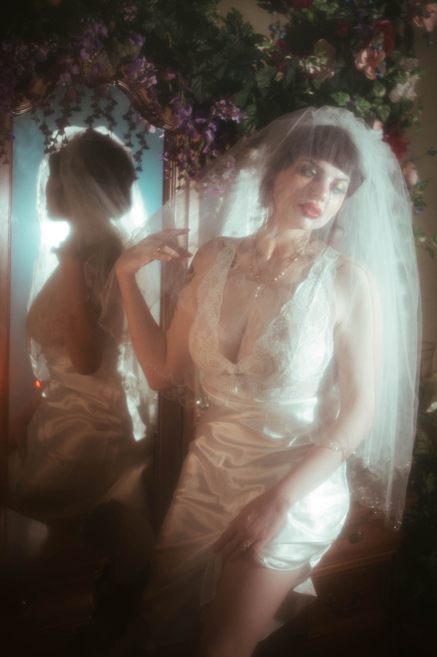A bride raises one hand to her veil while her reflection appears in an antique mirror beside a floral archway, soft fog machine boudoir photography haze glowing around her lace nightgown, Royal Lune Photo.
