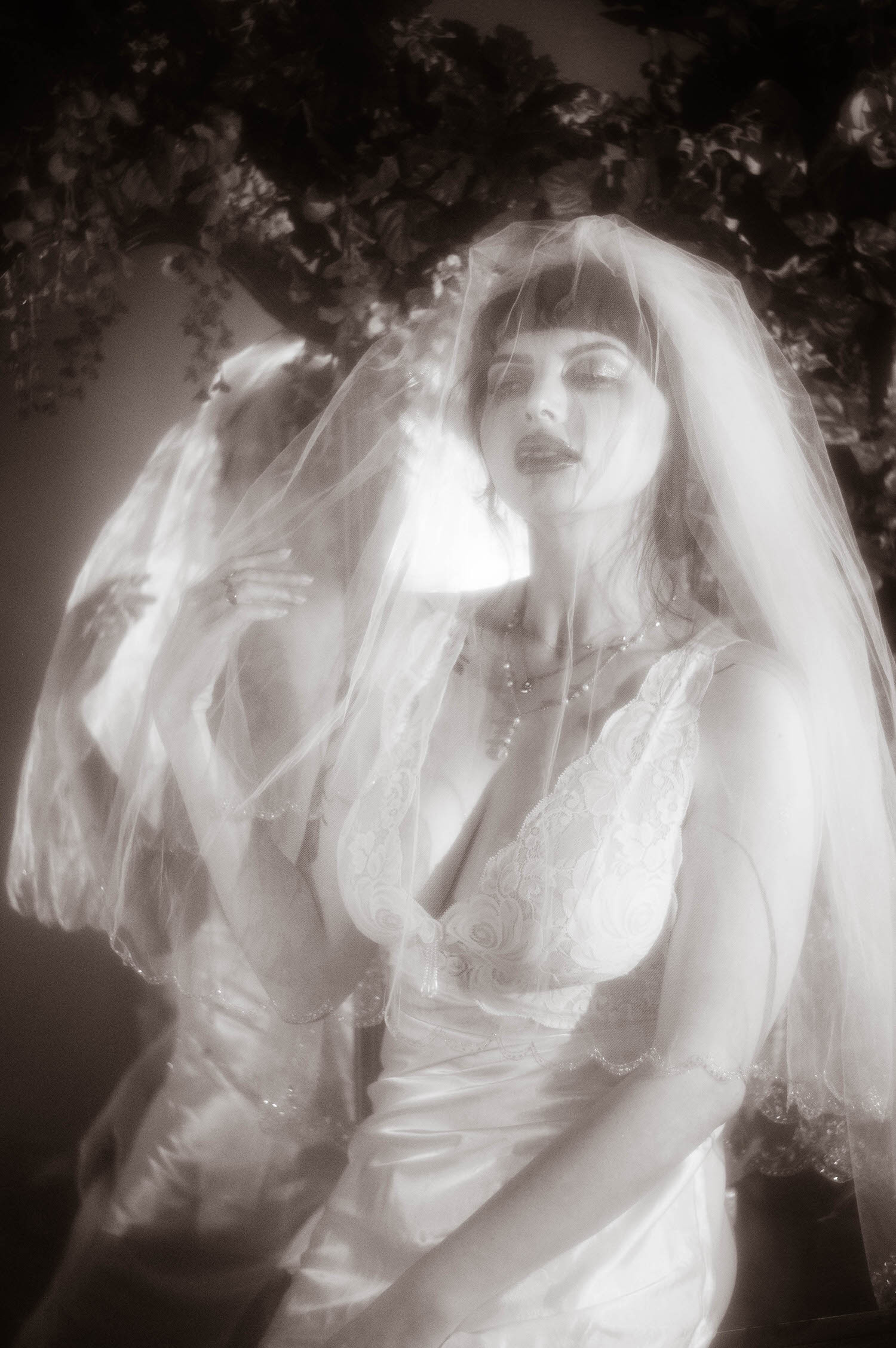 A black and white portrait shows a bride with one hand raised near her veil, soft tonal light tracing her lace nightgown and pearl necklace beneath a canopy of greenery at Royal Lune Studio, Royal Lune Photo.