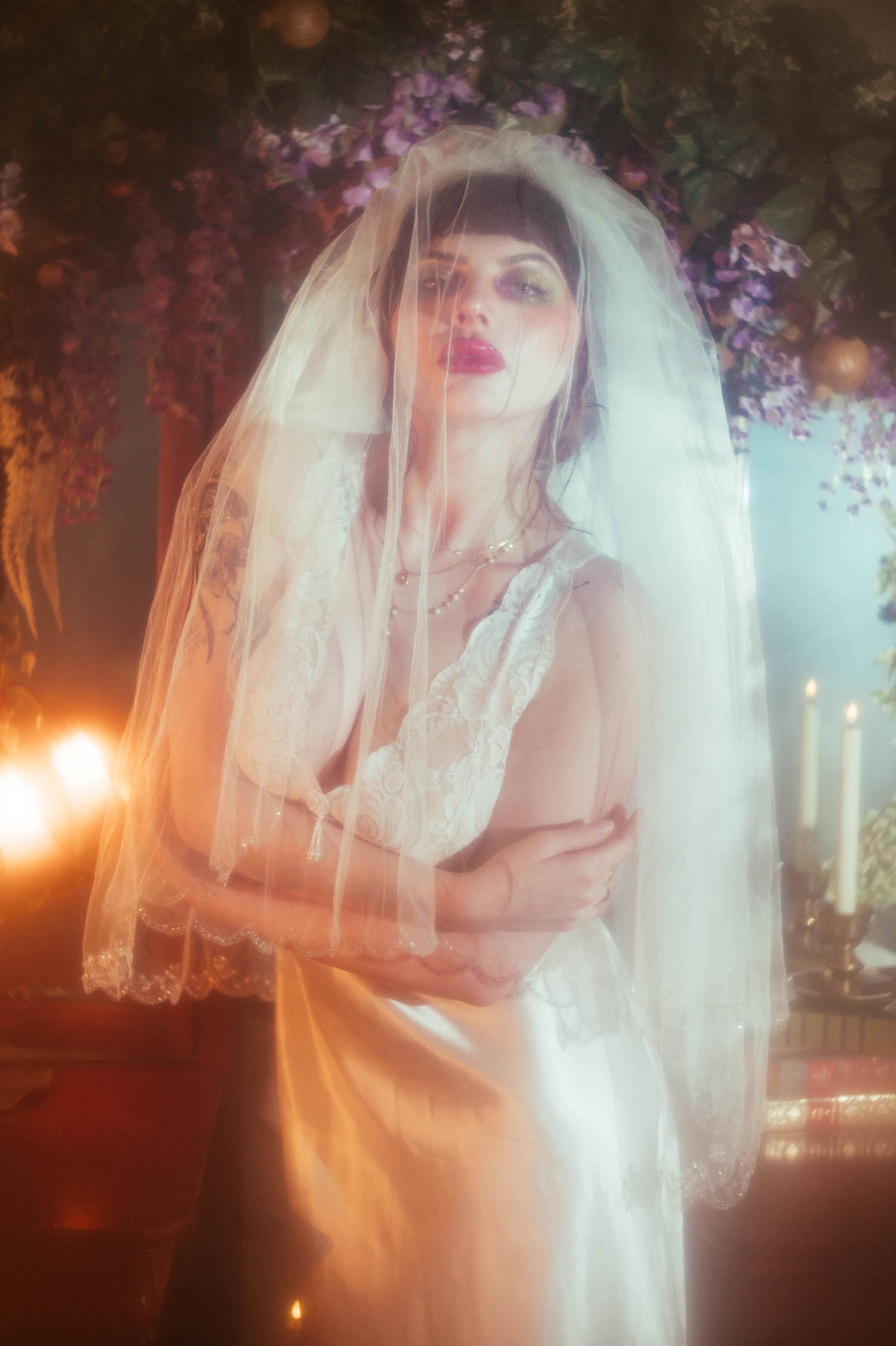 A bride stands with her arms crossed beneath a sheer veil, two lit candles glowing beside her as a lush floral archway and teal mirror fill the background of this moody boudoir lighting portrait, Royal Lune Photo.