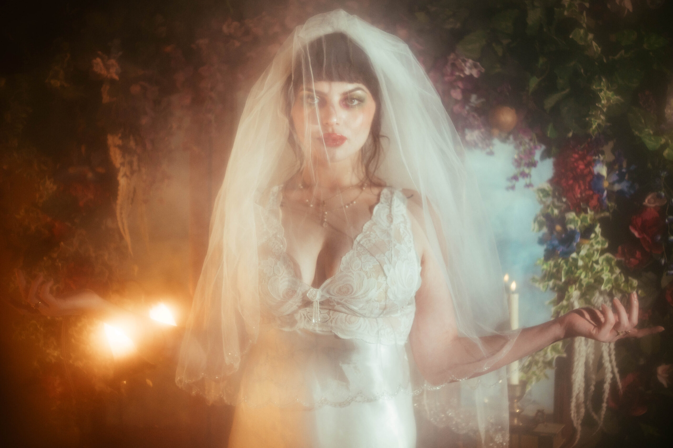 A bride stands with her arms open beneath a sheer veil, warm candlelight and a vivid floral archway framing her as a DFW bridal boudoir photographer captures this dreamy bridal boudoir photography moment, Royal Lune Photo.