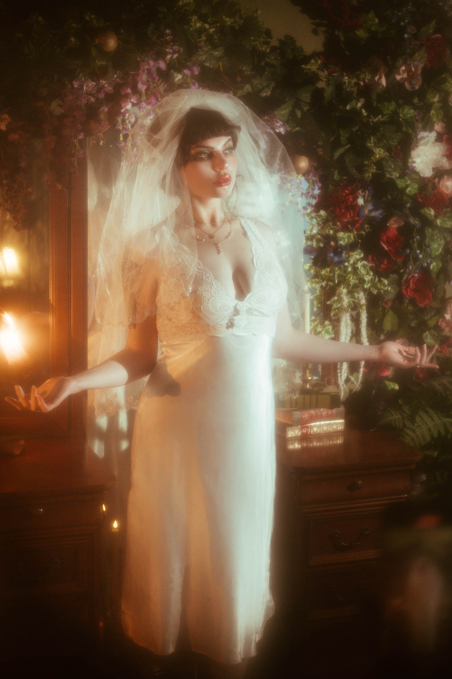 A bride in a vintage nightgown boudoir look stands with her arms outstretched, her sheer veil catching warm candlelight beside an antique wooden vanity and a lush floral archway inside Royal Lune's vintage photo studio, Royal Lune Photo.