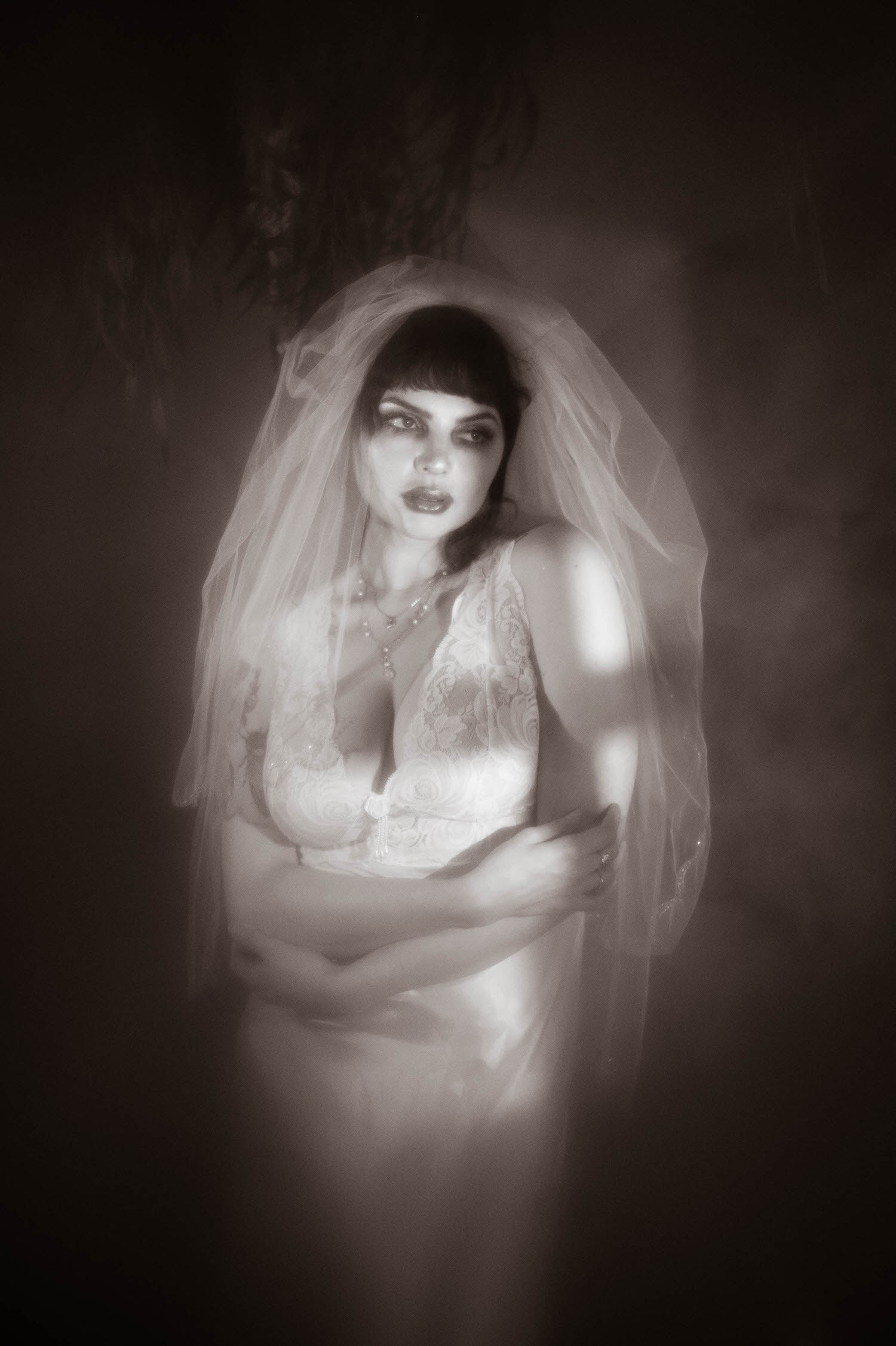 A black and white portrait shows a bride with her arms crossed beneath a sheer veil, soft diffused light glowing through heavy fog during this bridal boudoir photography session at Royal Lune Studio, Royal Lune Photo.