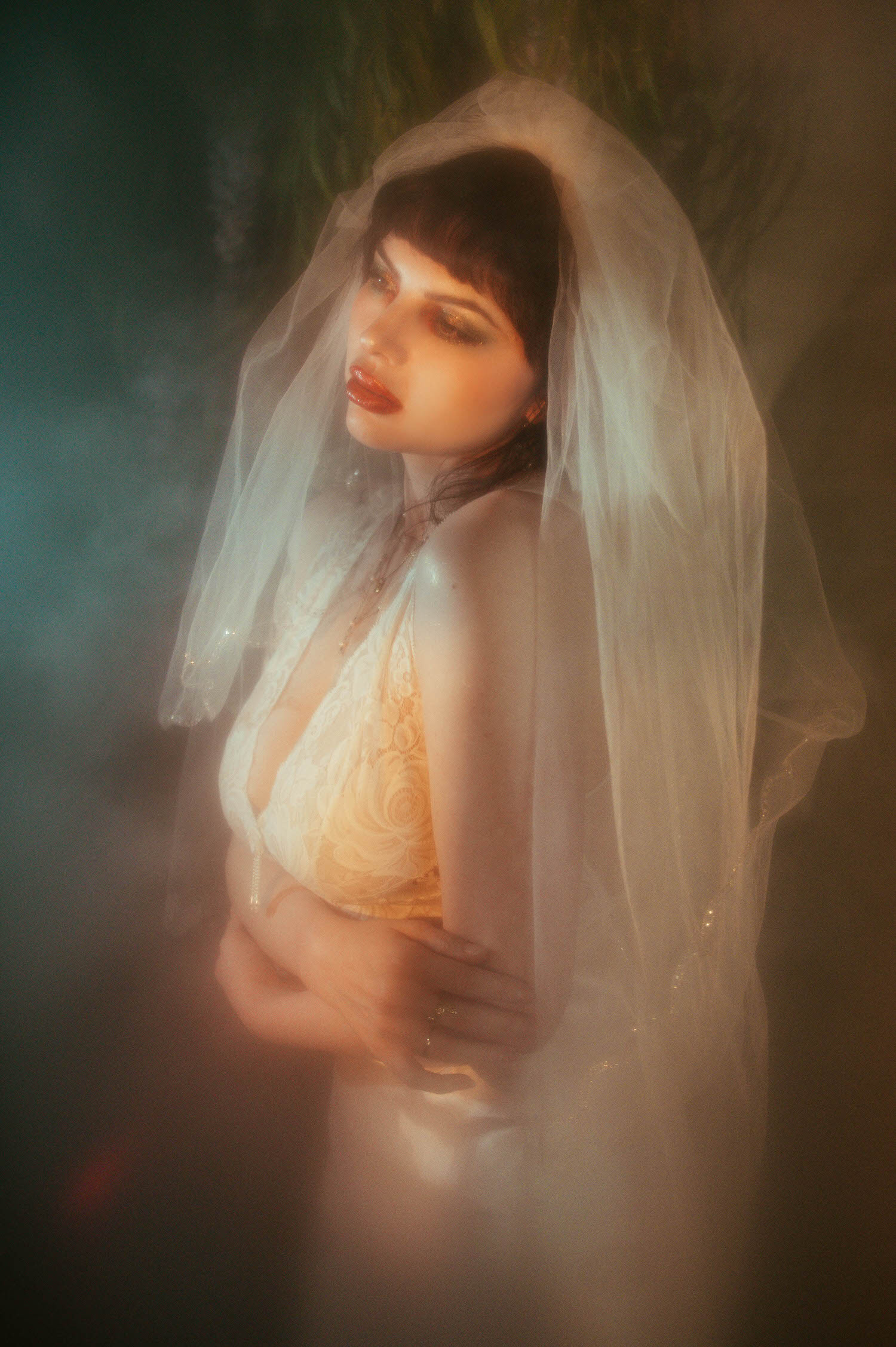 A bride in profile crosses her arms beneath a sheer veil, warm light catching her lace nightgown while a cool teal glow edges into the frame through dense fog machine boudoir photography haze, Royal Lune Photo.