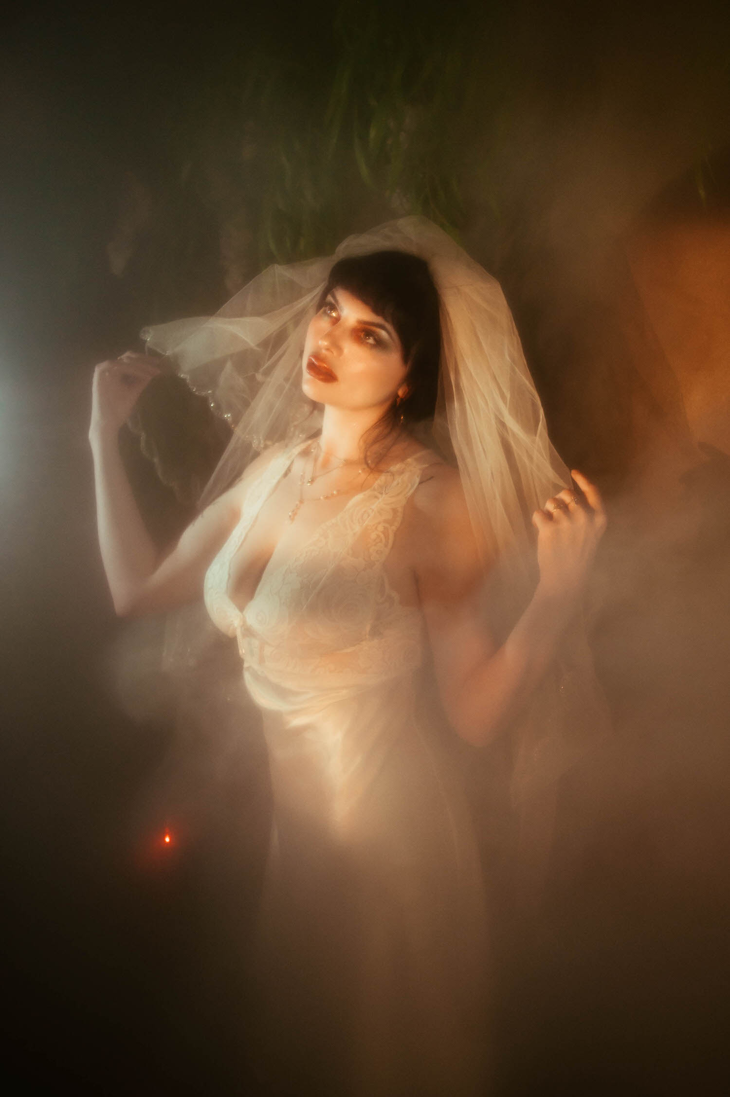 A bride raises both hands to her veil and looks upward, a small red light glowing in the fog machine boudoir photography haze behind her as a DFW bridal boudoir photographer captures the dramatic scene, Royal Lune Photo.