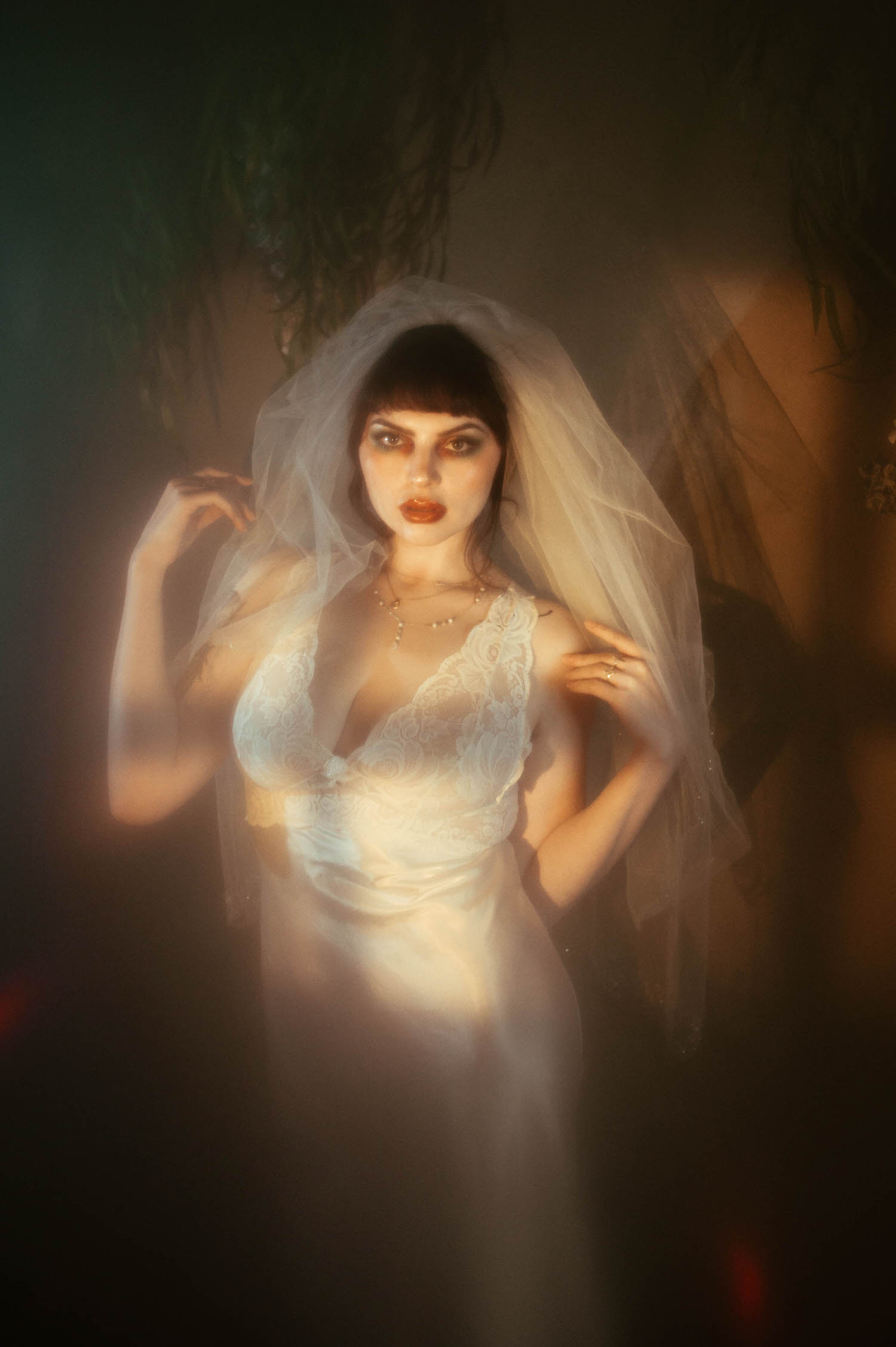A bride stands with one hand on her hip and the other holding her veil at her shoulder, warm moody boudoir lighting cutting through thick fog inside Royal Lune's vintage photo studio, Royal Lune Photo.