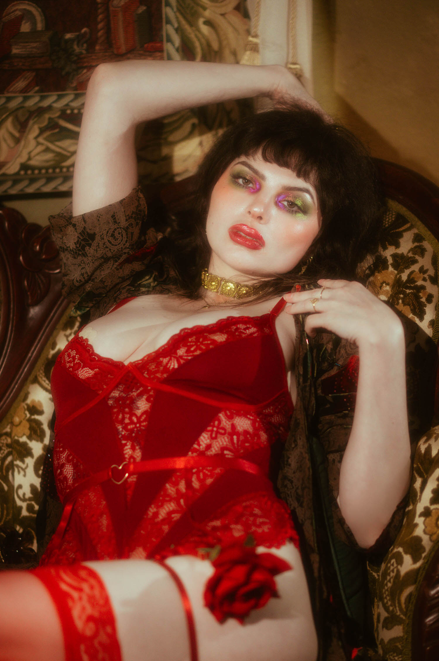 A woman reclines against a floral tapestry sofa in red lace lingerie with scalloped trim and a heart shaped buckle detail, a red rose resting near her thigh in this red rose boudoir photoshoot, Royal Lune Photo.