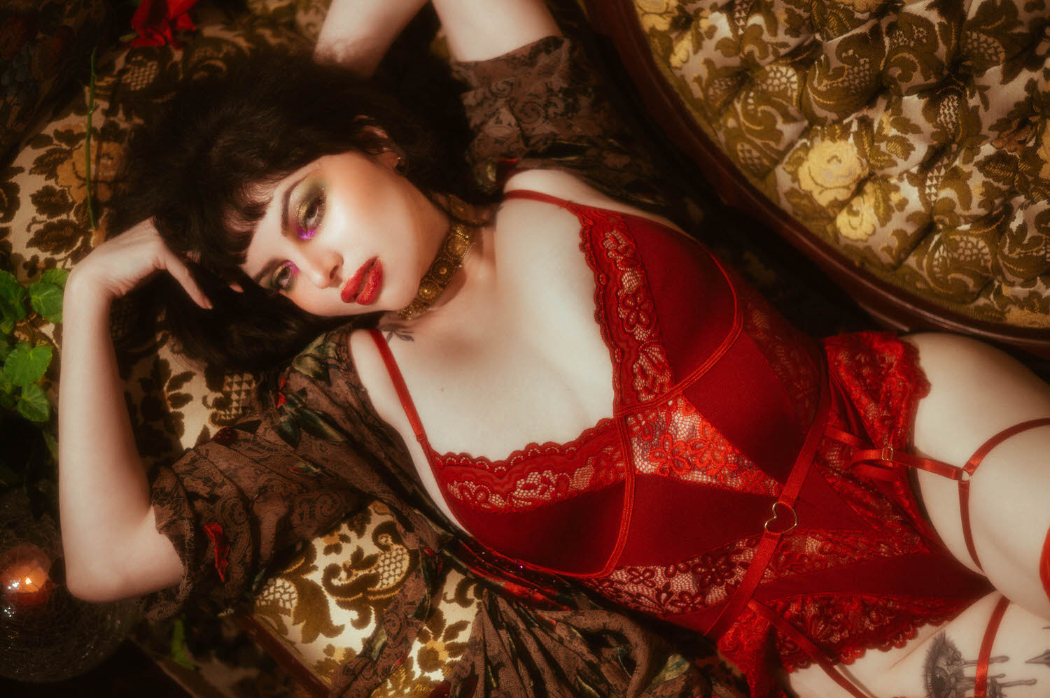 A woman rests her head against a floral tapestry sofa in red lace lingerie with a heart shaped buckle detail, wearing a gold beaded choker and the whimsigoth silk robe in this fine art boudoir photography session, Royal Lune Photo.