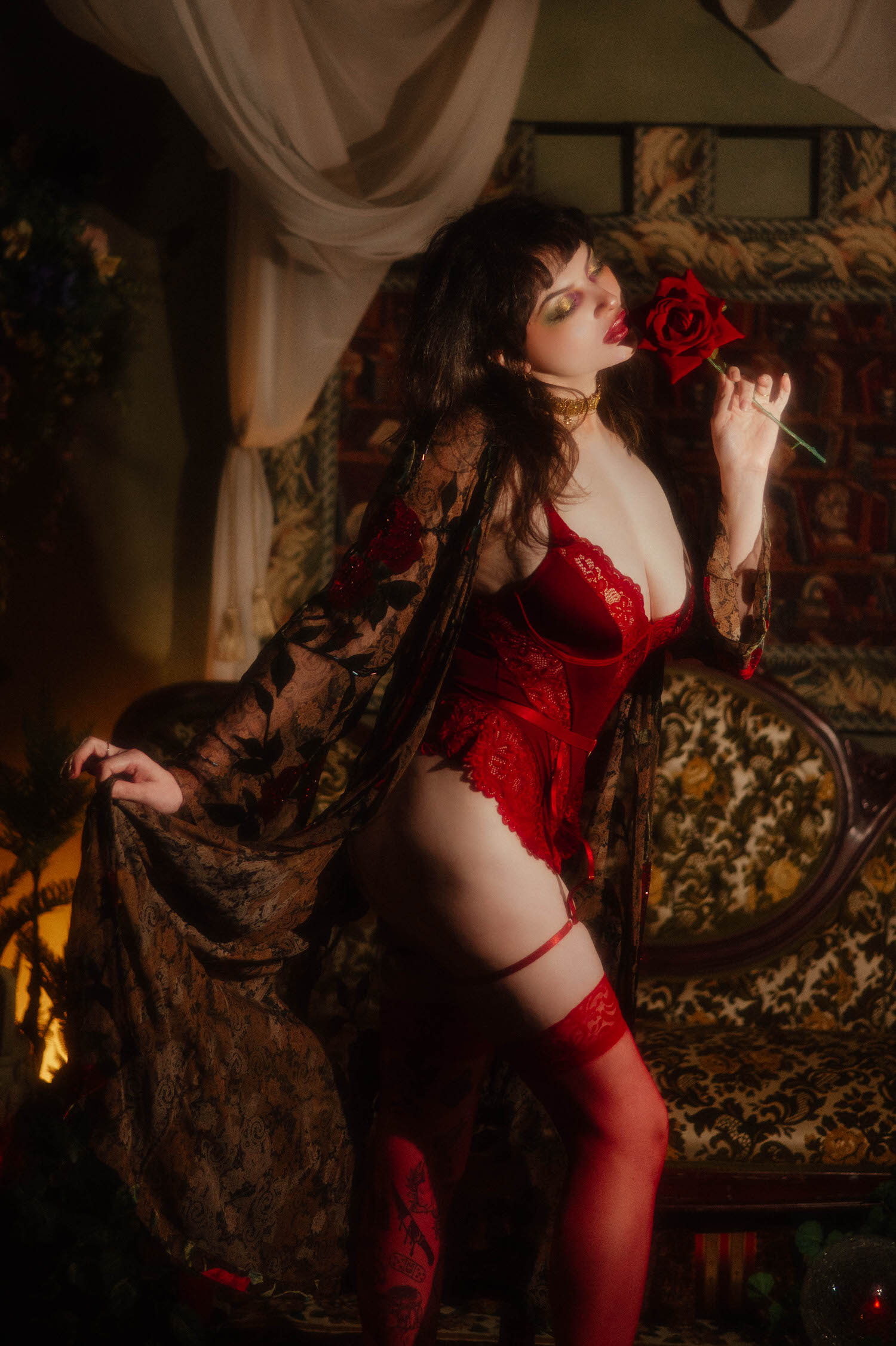 A woman poses in red lace lingerie and a whimsigoth silk robe with a beaded rose design, holding a red rose to her lips beside sheer cream curtains and an antique tufted sofa in this red rose boudoir photoshoot at Royal Lune Studio, Royal Lune Photo.