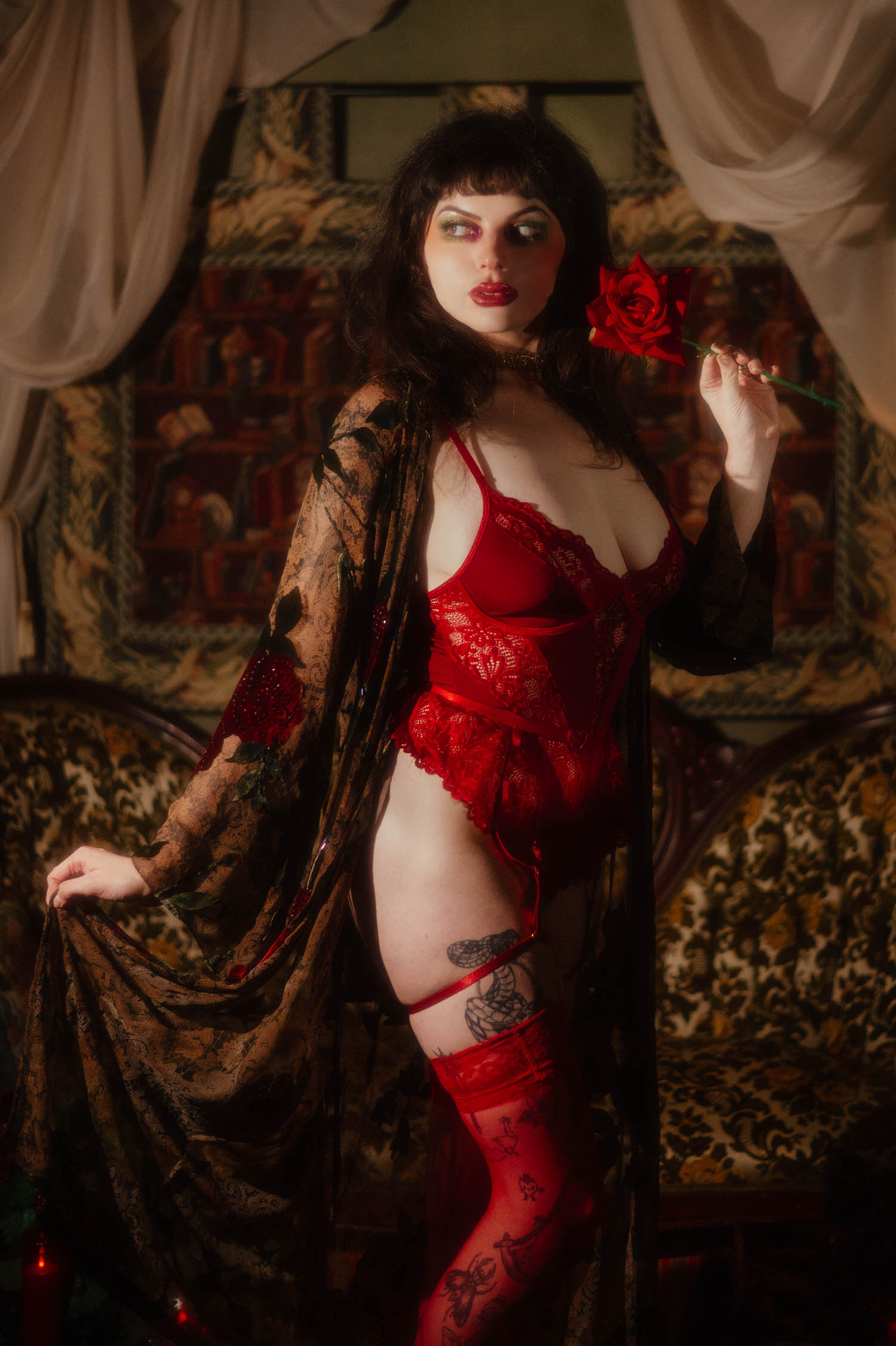 A woman stands in red lace lingerie and sheer red thigh high stockings, holding a red rose and gathering the whimsigoth silk robe in one hand against a library tapestry backdrop, Royal Lune Photo.
