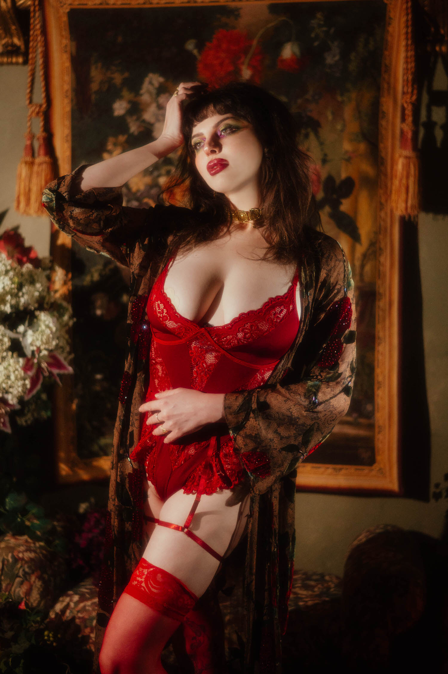 A woman stands wearing the whimsigoth silk robe over red lace lingerie with sheer red thigh high stockings, styled against a large floral painting with gold tasseled drapery cords in this red rose boudoir photoshoot, Royal Lune Photo.