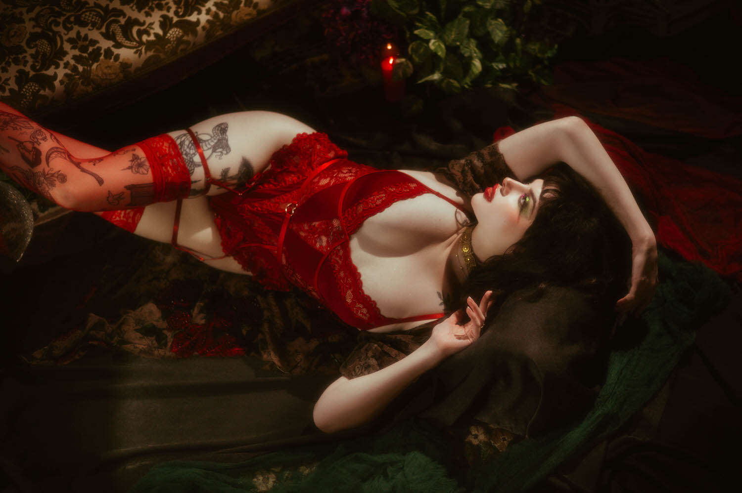 A woman lies back with one arm raised, wearing red lace lingerie with sheer red thigh high stockings and a gold beaded choker, the whimsigoth robe sleeve visible beside her on a dark floral fabric backdrop, Royal Lune Photo.