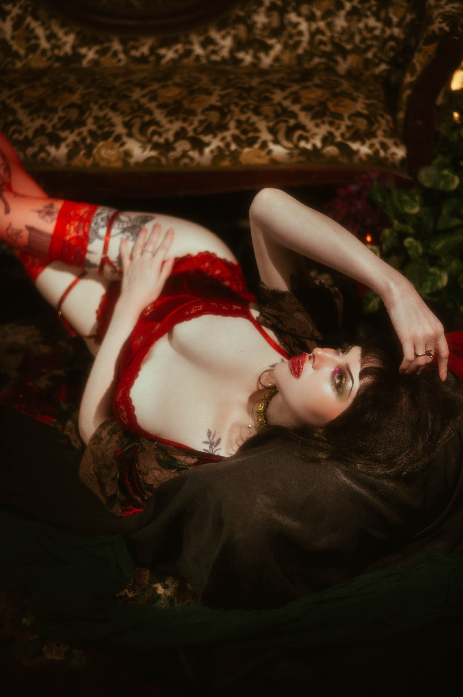 A woman reclines with one leg raised on a vintage tapestry sofa, wearing red lace lingerie, sheer red thigh high stockings, and the whimsigoth silk robe, a floral tattoo visible on her chest in this fine art boudoir photography session, Royal Lune Photo.