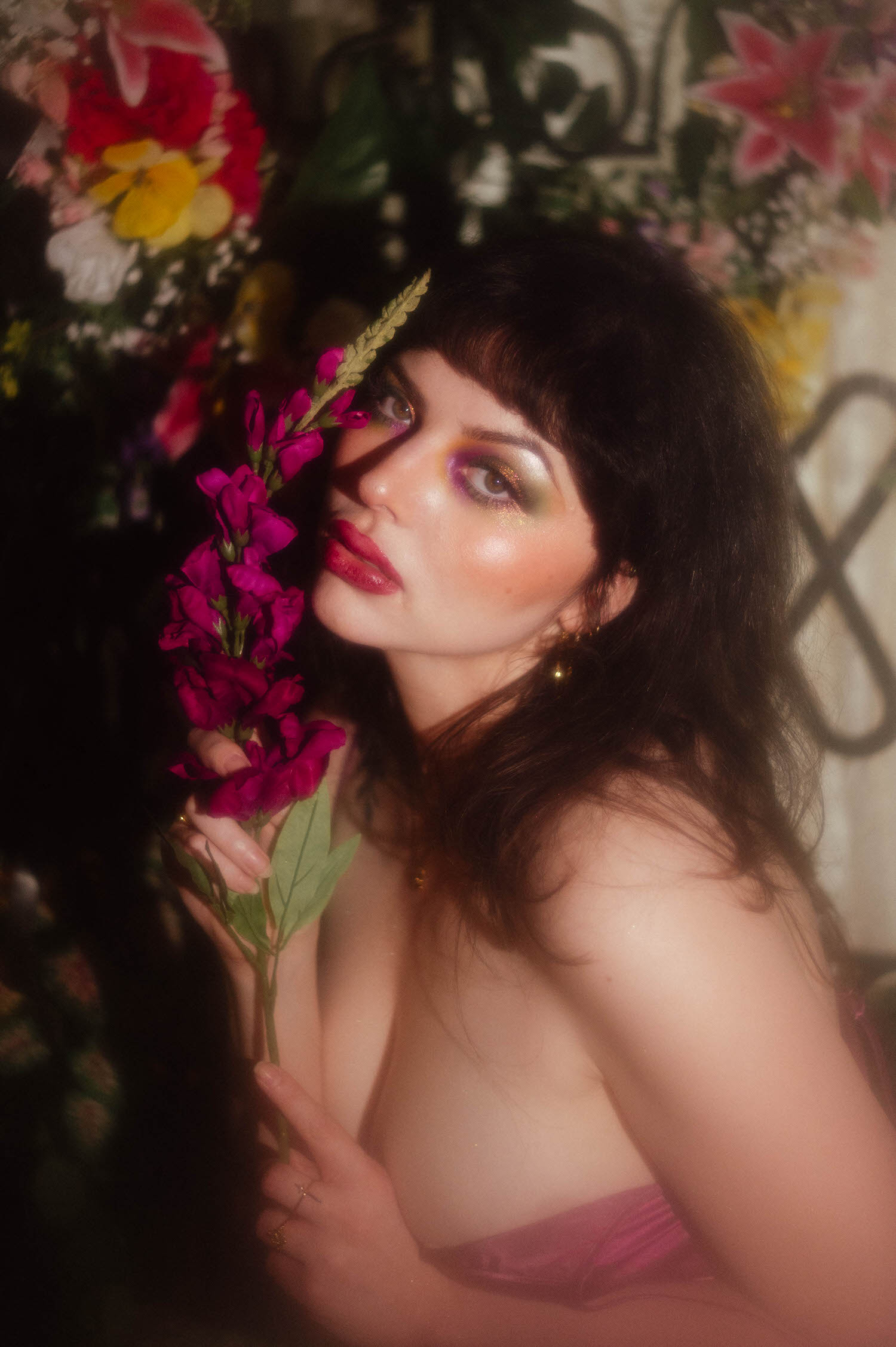 A bride wearing a magenta satin slip dress poses with fuchsia florals tucked into her hair and fine line tattoos visible along her arm, set against a colorful floral arrangement and iron gate during this bridal boudoir photography session at Royal Lune Studio, Royal Lune Photo.