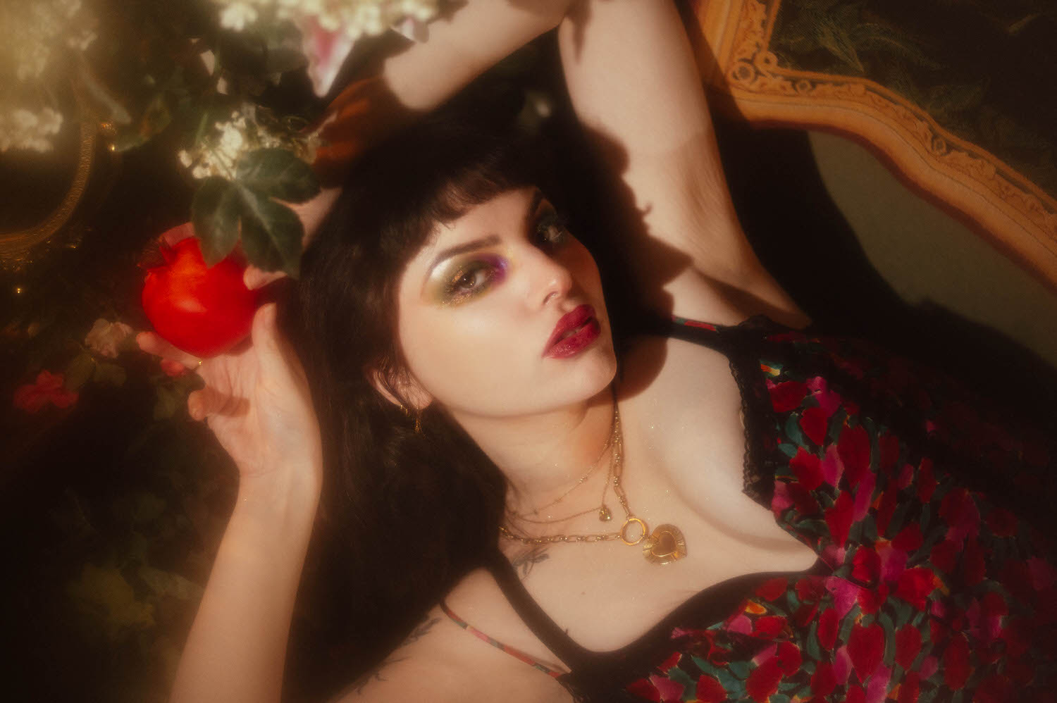A close up portrait shows a woman holding a red pomegranate near her face, wearing a gold heart pendant necklace and a floral print lingerie top, with gold and purple eye makeup in this fine art boudoir photography session, Royal Lune Photo.