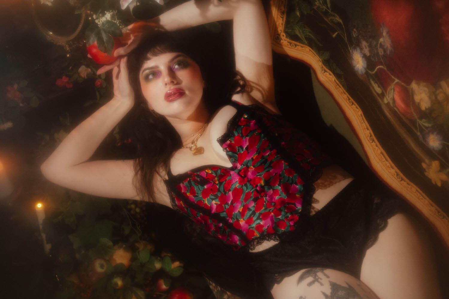  A woman lies back on a floral tapestry backdrop in a vintage two piece lingerie set with a bold red, pink, and green floral print, reaching up toward a pomegranate prop in this red rose boudoir photoshoot at Royal Lune Studio, Royal Lune Photo.