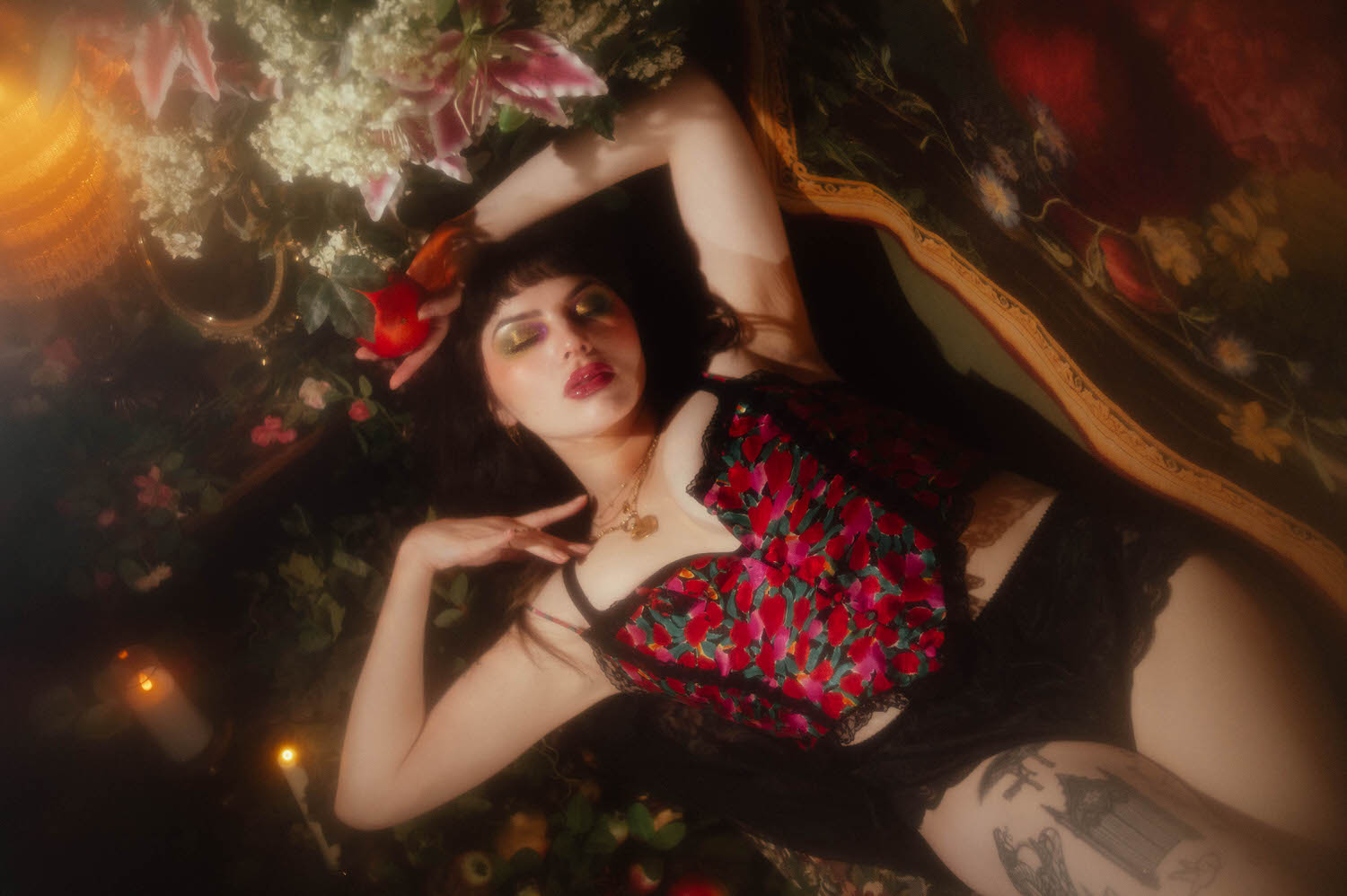 A woman lies among pink stargazer lilies and baby's breath holding a pomegranate near her head, wearing a vintage floral lingerie set and layered gold necklaces, lit by warm candlelight in this dark romantic boudoir photography session, Royal Lune Photo.