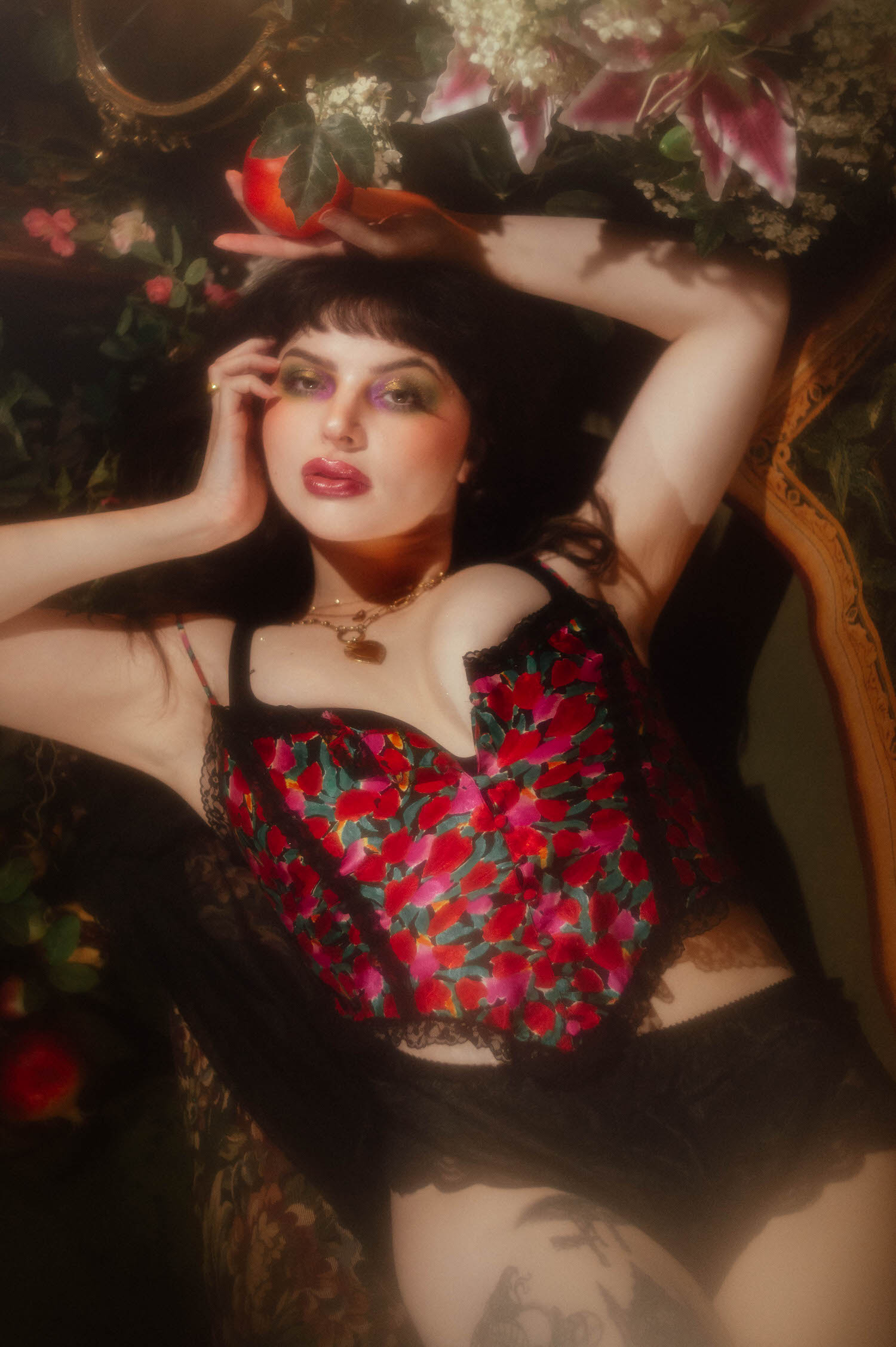 A woman rests her head in her hand while holding a pomegranate above her in a vintage lingerie boudoir photoshoot, wearing a gold pendant necklace and a red, pink, and green floral bralette, styled with an antique mirror in the background, Royal Lune Photo.